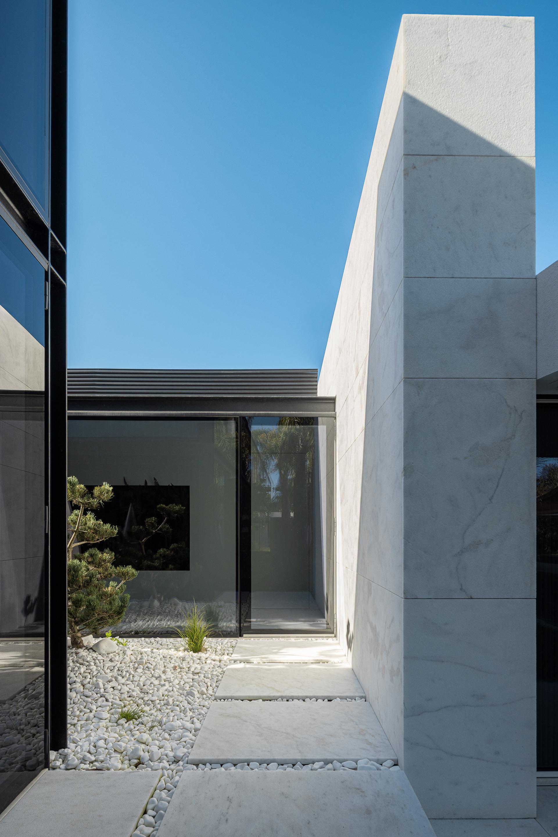 Corredor exterior entre volumes, com pilar em mármore branco, paredes envidraçadas e pavimento de seixos — RiscoWhite arquitectura Risco Singular – Arquitectura, Lda. fotografia Ivo Tavares