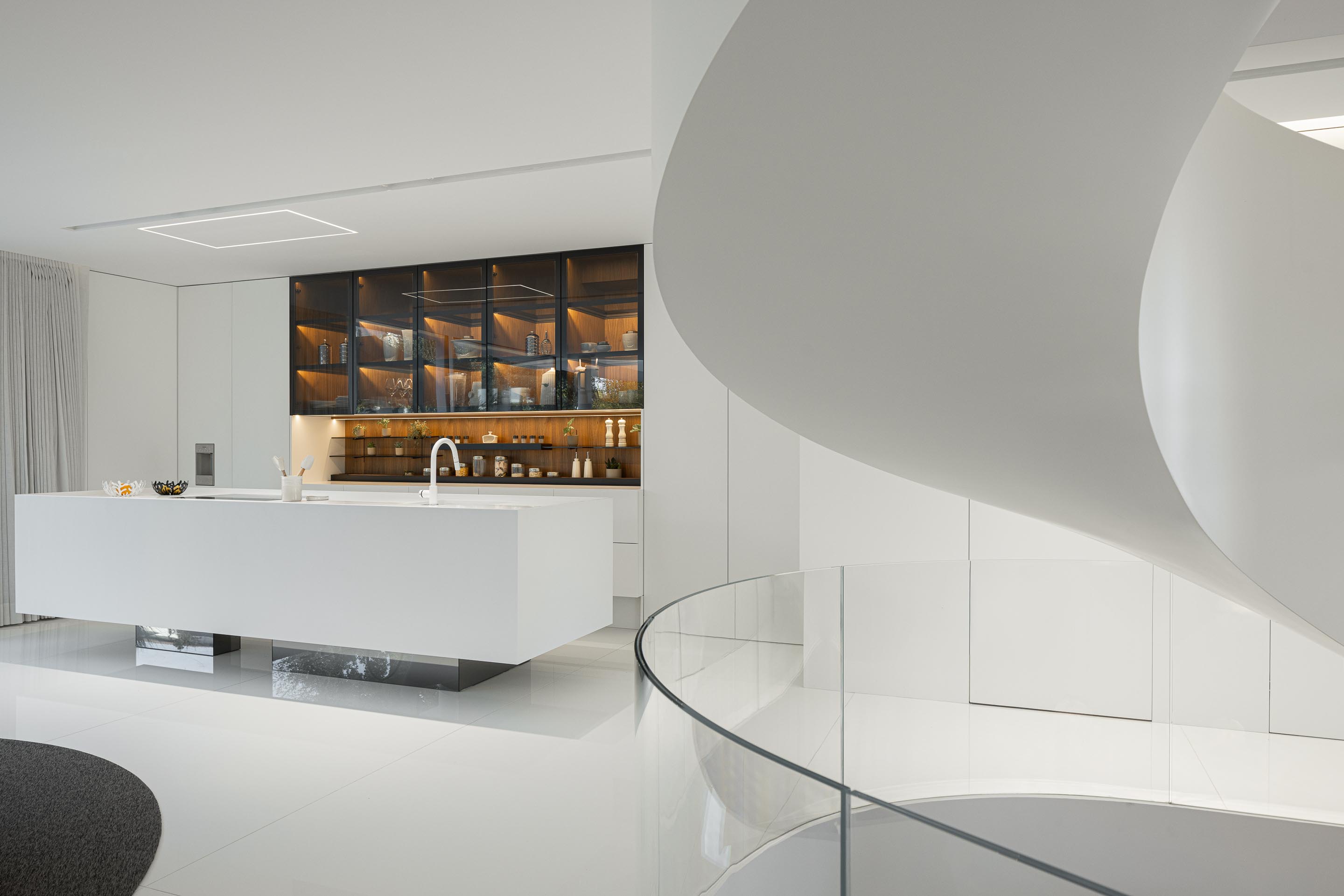 Cozinha com ilha central branca e armários superiores iluminados, adjacente à escada em caracol — RiscoWhite arquitectura Risco Singular – Arquitectura, Lda. fotografia Ivo Tavares