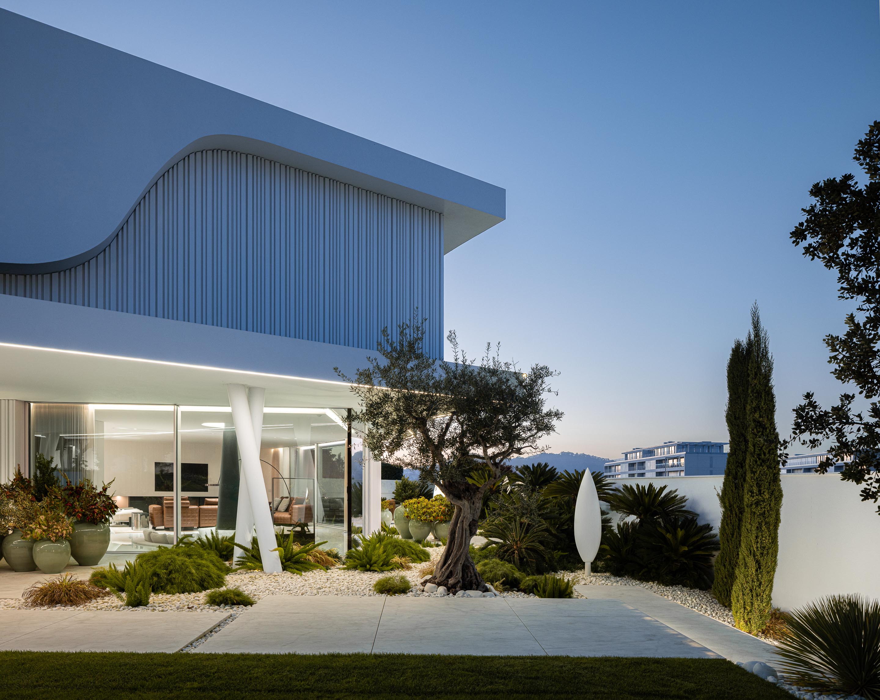Vista lateral da moradia ao anoitecer, com formas curvas, grandes vãos envidraçados e jardim iluminado — RiscoWhite arquitectura Risco Singular – Arquitectura, Lda. fotografia Ivo Tavares