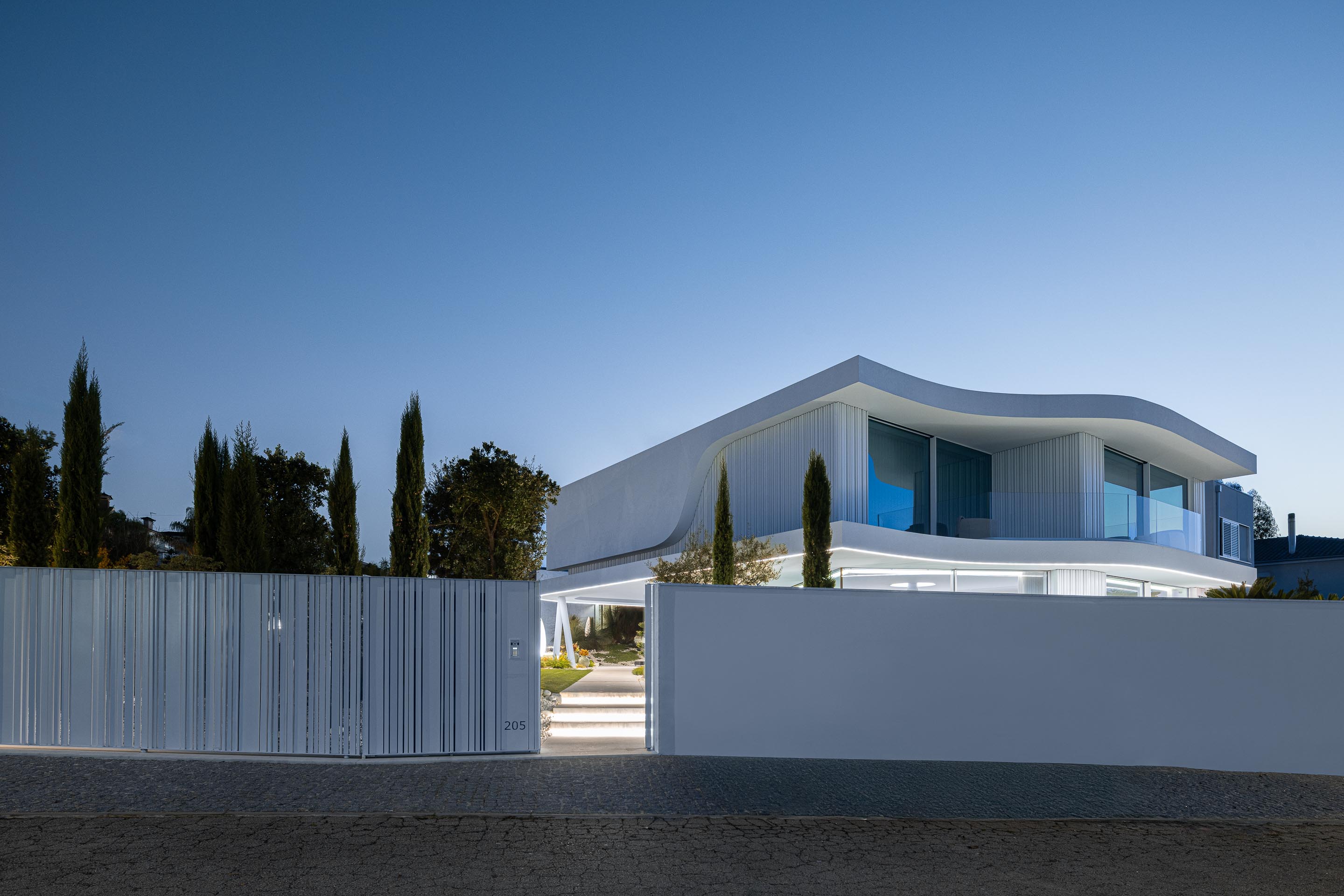Entrada da propriedade ao anoitecer, com portão de ripado vertical branco e moradia iluminada ao fundo — RiscoWhite arquitectura Risco Singular – Arquitectura, Lda. fotografia Ivo Tavares