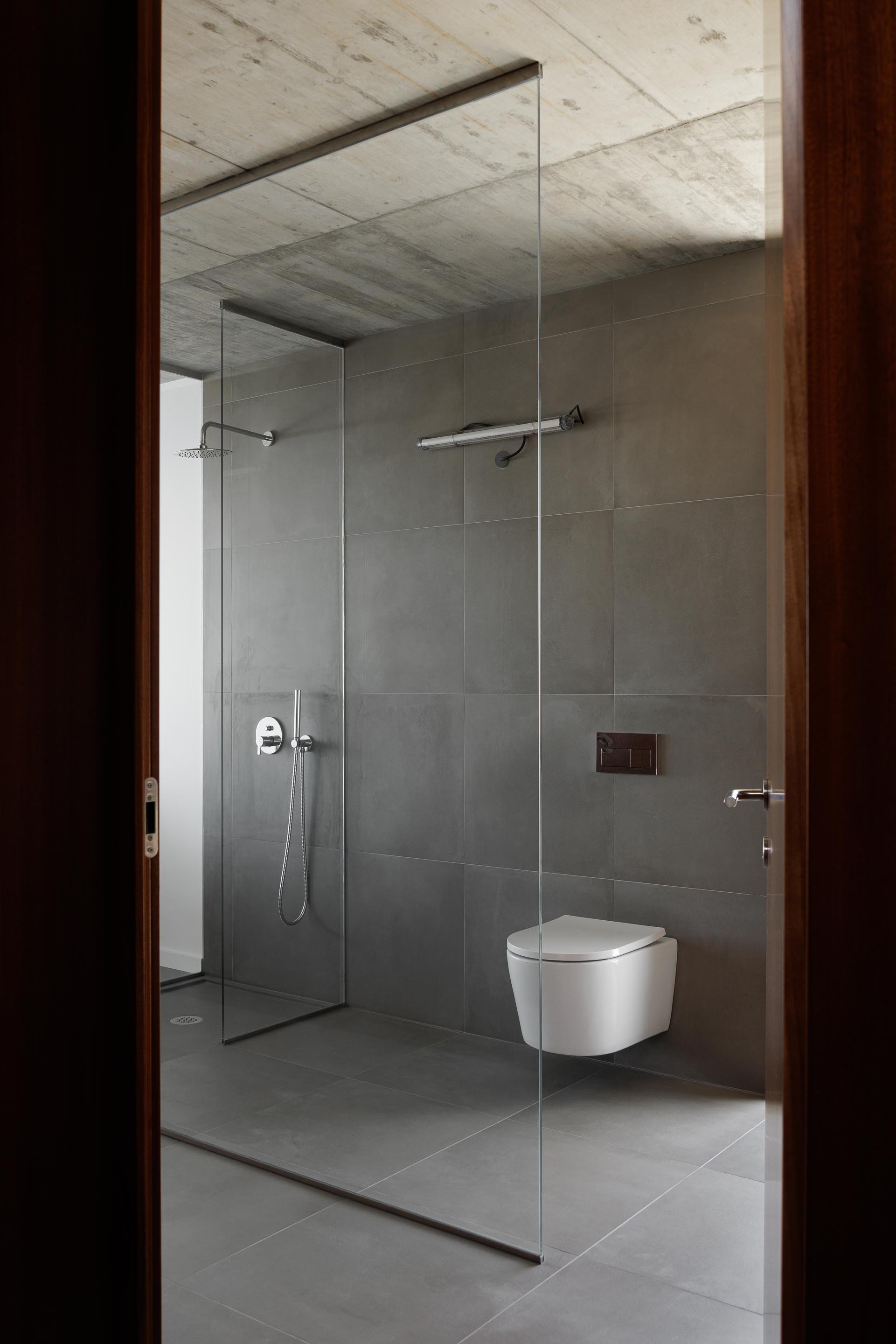 Casa de banho com pavimento e paredes em cerâmica cinzenta, duche em vidro estrutural e teto em betão aparente — CASA COQUEIRA arquitectura Tiago Sousa Arquitecto fotografia Ivo Tavares