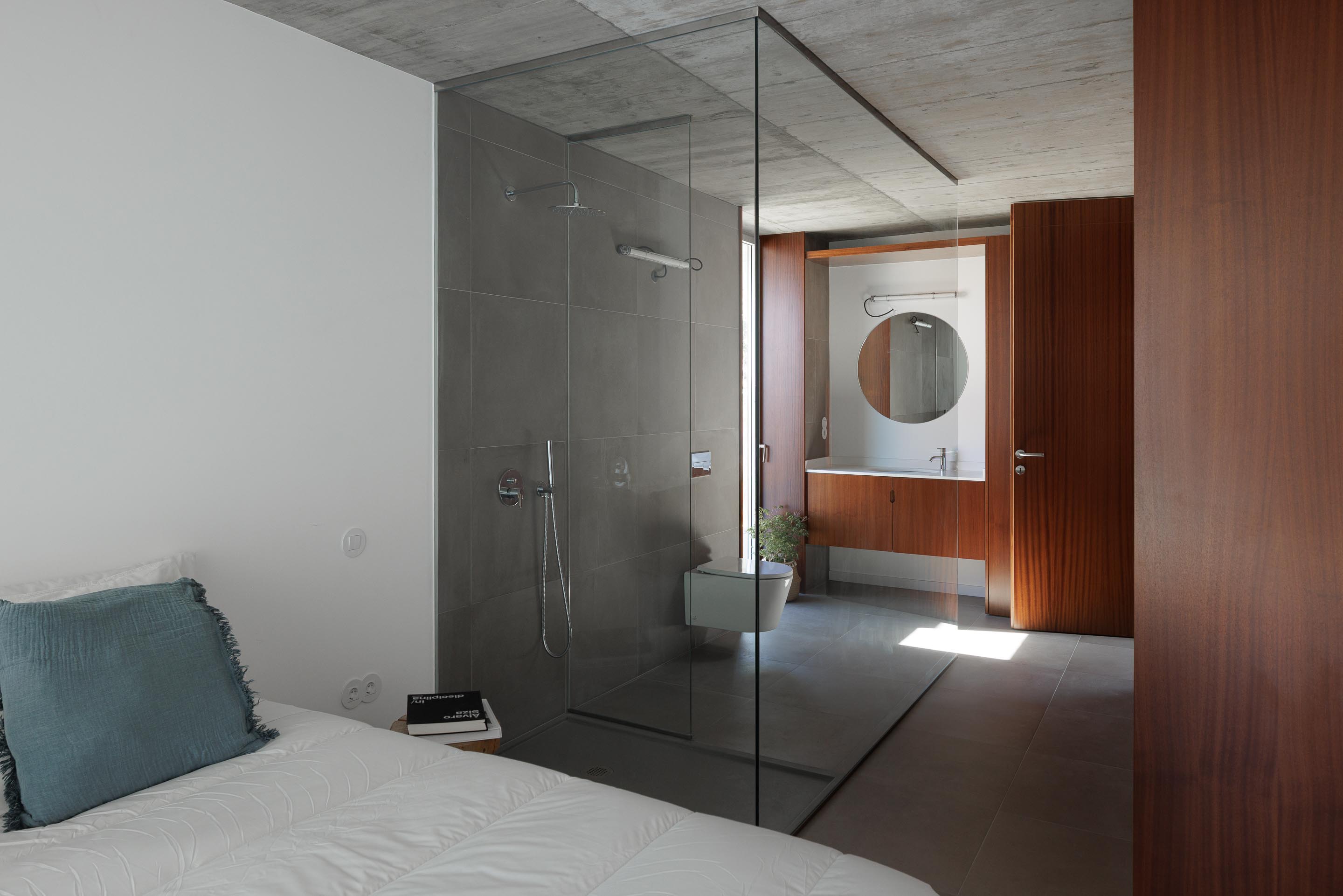 Quarto com cama branca, casa de banho integrada com duche em vidro e lavatório em madeira escura. Teto em betão aparente — CASA COQUEIRA arquitectura Tiago Sousa Arquitecto fotografia Ivo Tavares