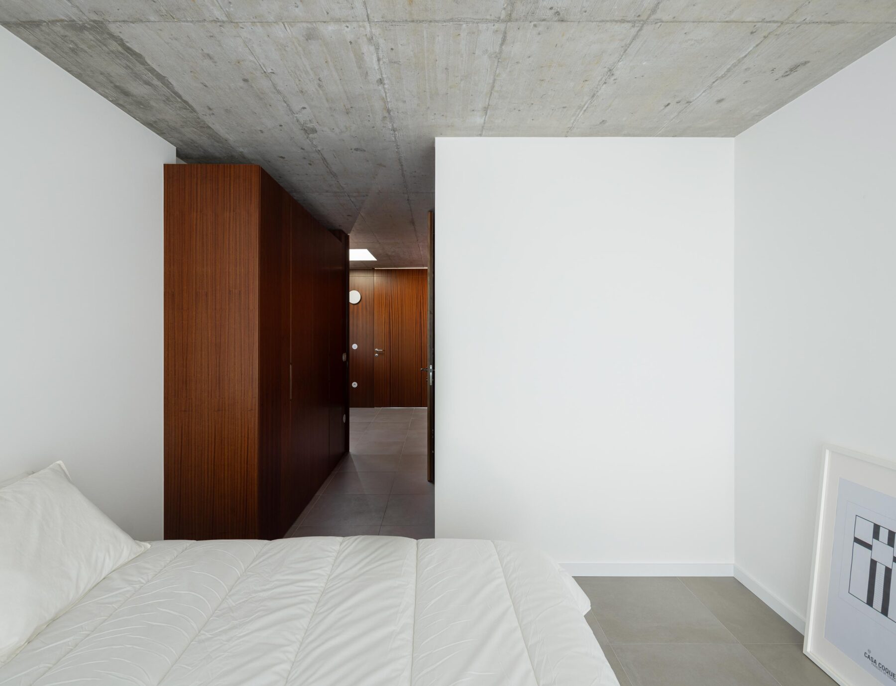 Quarto com paredes brancas e pavimento cinzento, armários em madeira escura e teto em betão aparente. Corredor visível — CASA COQUEIRA arquitectura Tiago Sousa Arquitecto fotografia Ivo Tavares