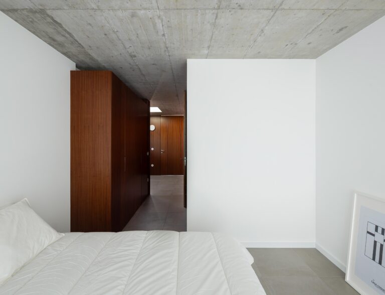 Quarto com paredes brancas e pavimento cinzento, armários em madeira escura e teto em betão aparente. Corredor visível — CASA COQUEIRA arquitectura Tiago Sousa Arquitecto fotografia Ivo Tavares