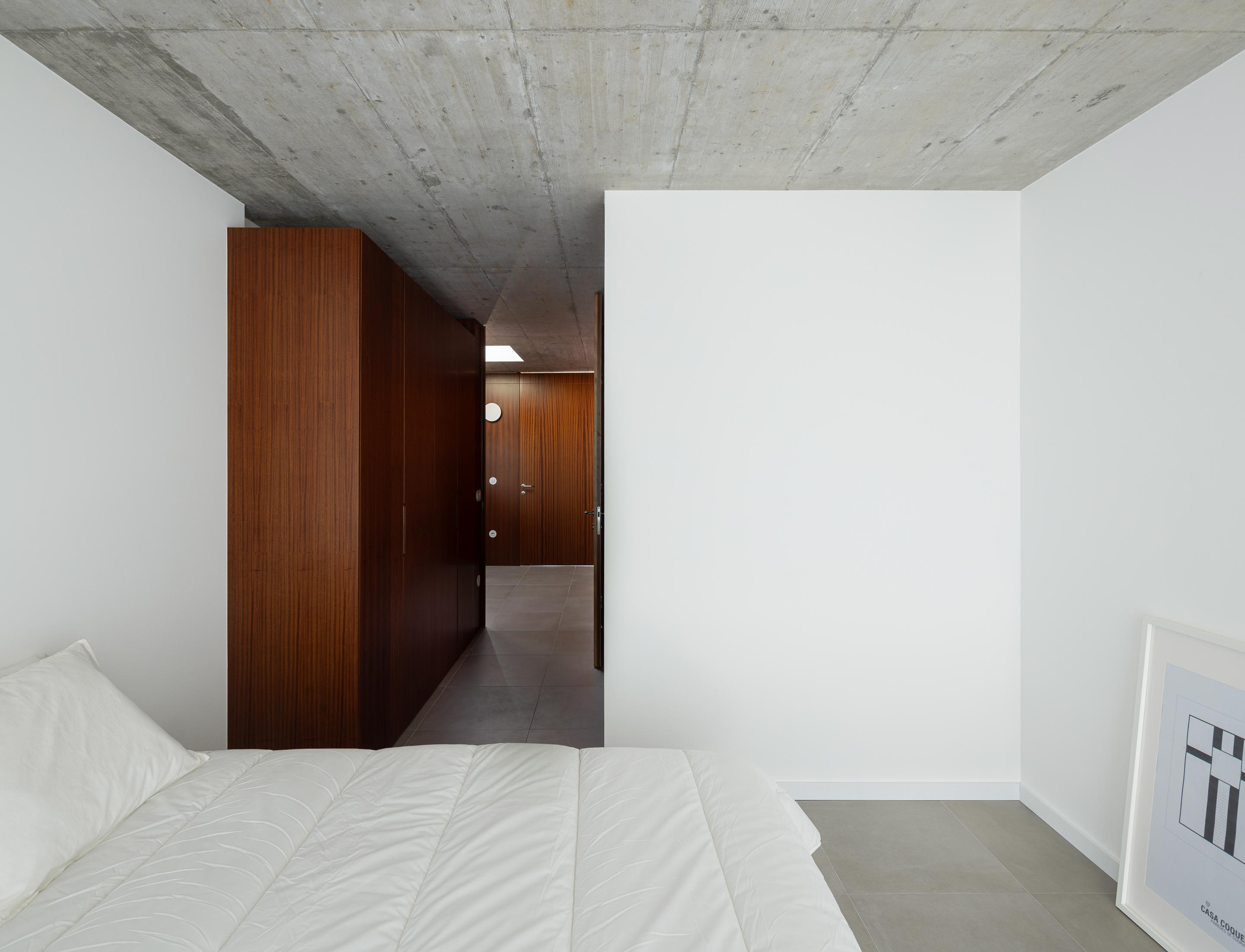 Quarto com paredes brancas e pavimento cinzento, armários em madeira escura e teto em betão aparente. Corredor visível — CASA COQUEIRA arquitectura Tiago Sousa Arquitecto fotografia Ivo Tavares