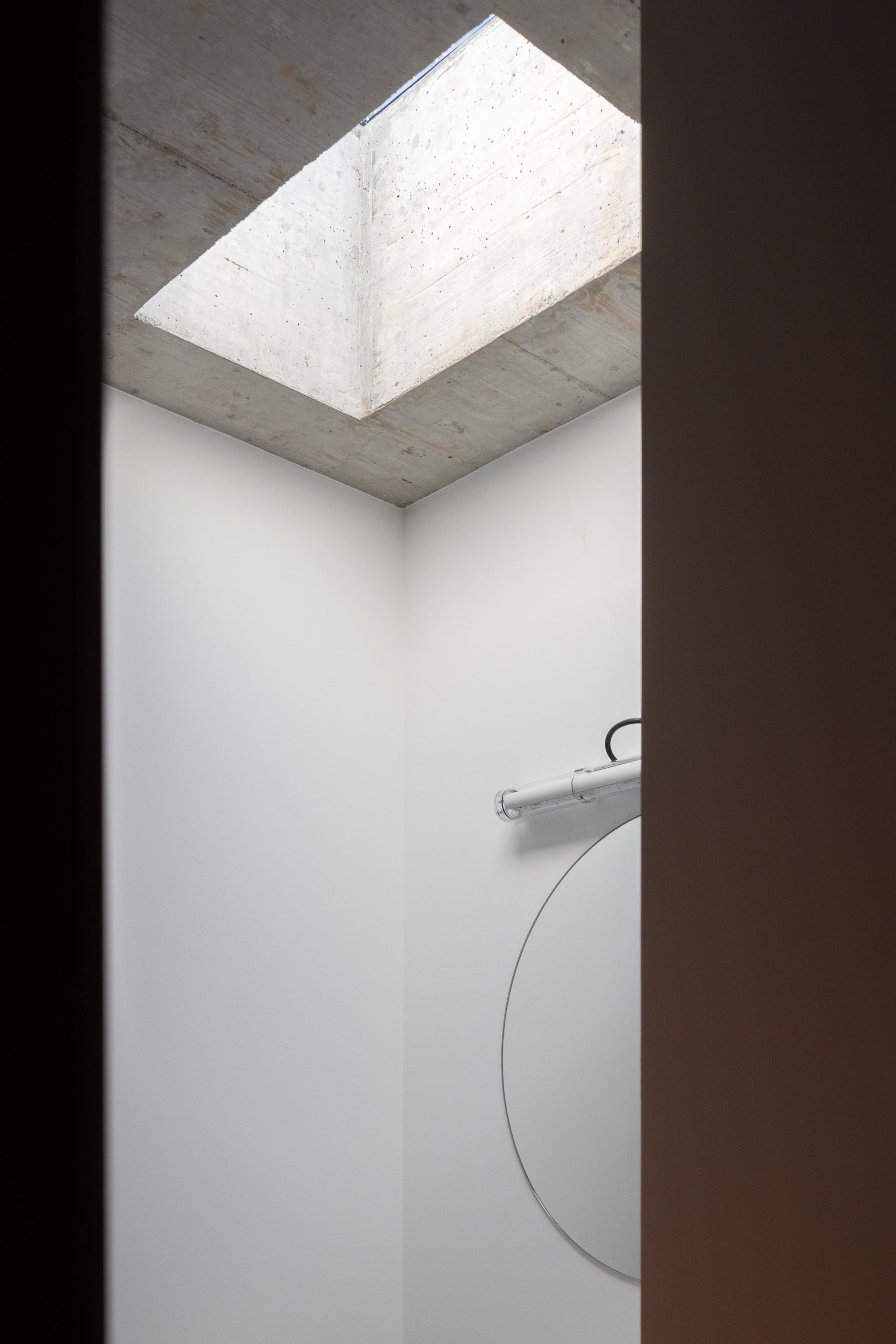 Pequena casa de banho com paredes brancas, espelho redondo e iluminação zenital através de claraboia retangular. Teto em betão aparente — CASA COQUEIRA arquitectura Tiago Sousa Arquitecto fotografia Ivo Tavares