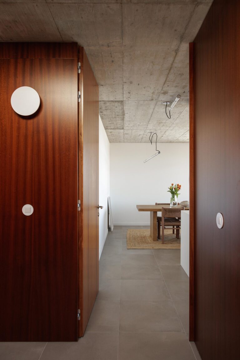 Corredor com pavimento cerâmico cinzento e teto em betão aparente, conduzindo à sala de jantar com mobiliário em madeira — CASA COQUEIRA arquitectura Tiago Sousa Arquitecto fotografia Ivo Tavares