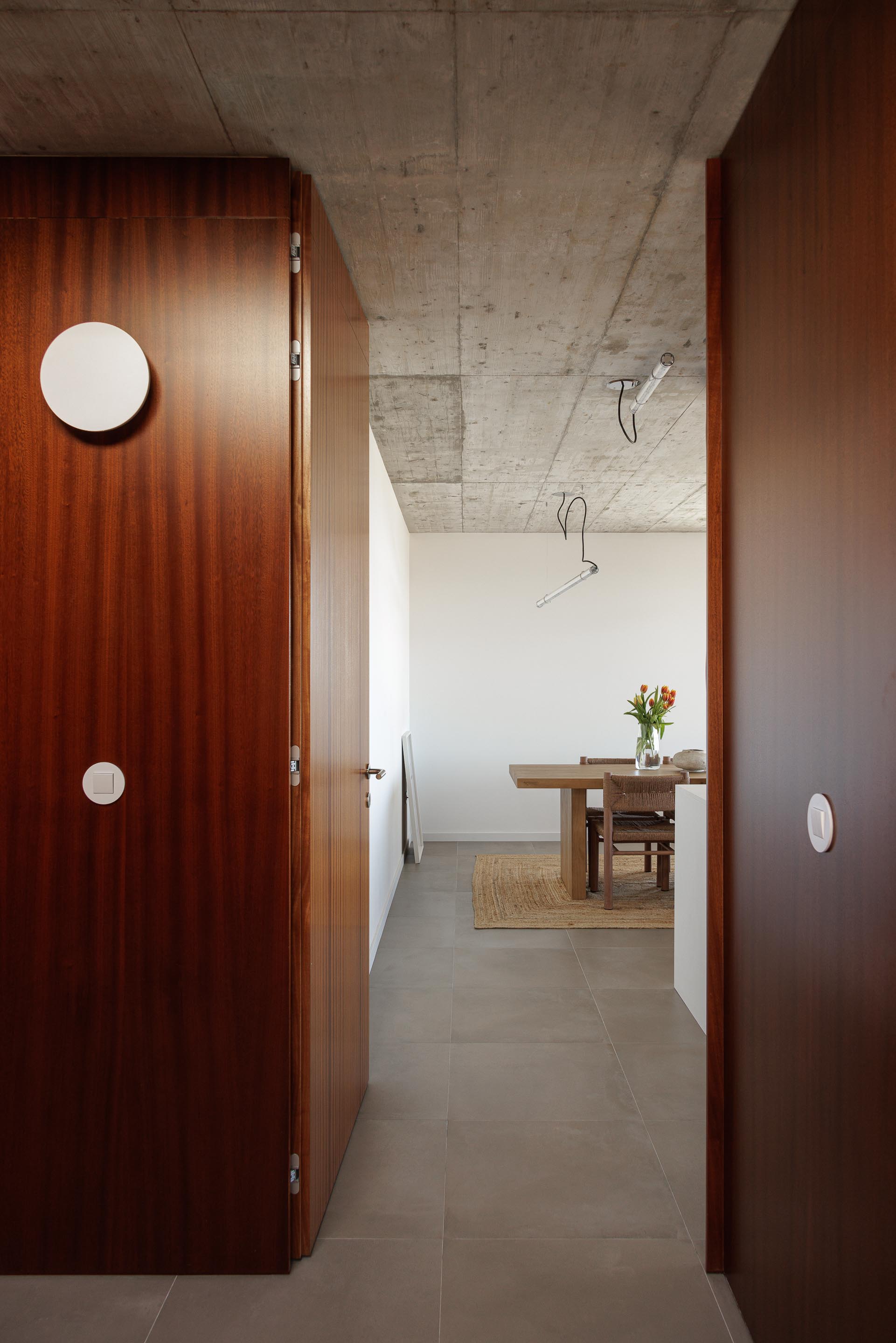 Corredor com pavimento cerâmico cinzento e teto em betão aparente, conduzindo à sala de jantar com mobiliário em madeira — CASA COQUEIRA arquitectura Tiago Sousa Arquitecto fotografia Ivo Tavares