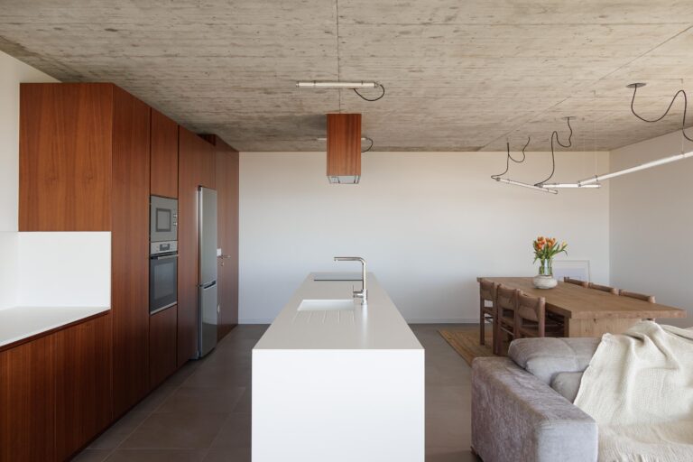 Cozinha e sala de jantar em planta aberta com teto em betão aparente, armários em madeira escura e ilha branca — CASA COQUEIRA arquitectura Tiago Sousa Arquitecto fotografia Ivo Tavares