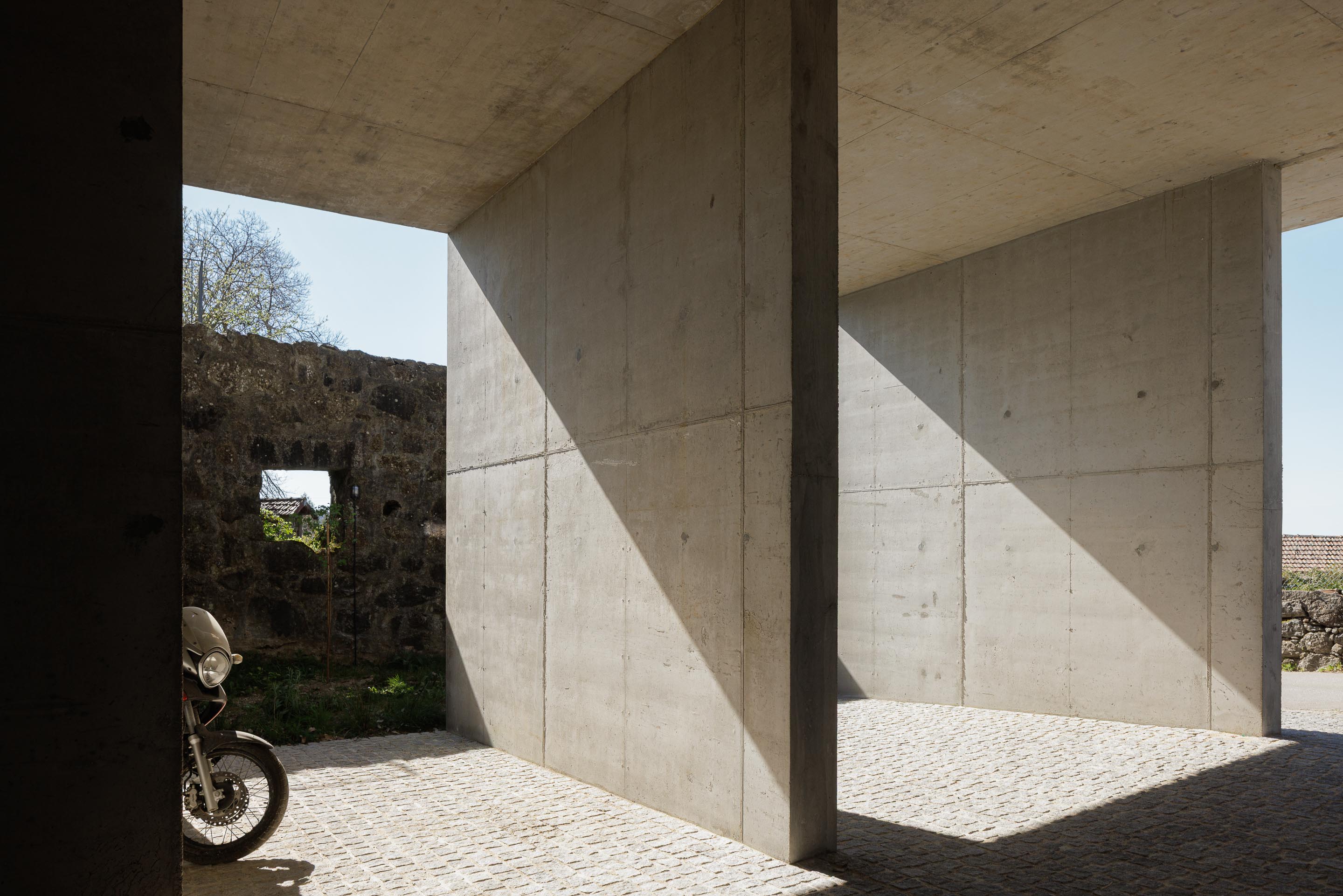 Espaço exterior coberto em betão aparente, com pavimento em calçada, pilares robustos e muro de pedra tradicional — CASA COQUEIRA arquitectura Tiago Sousa Arquitecto fotografia Ivo Tavares