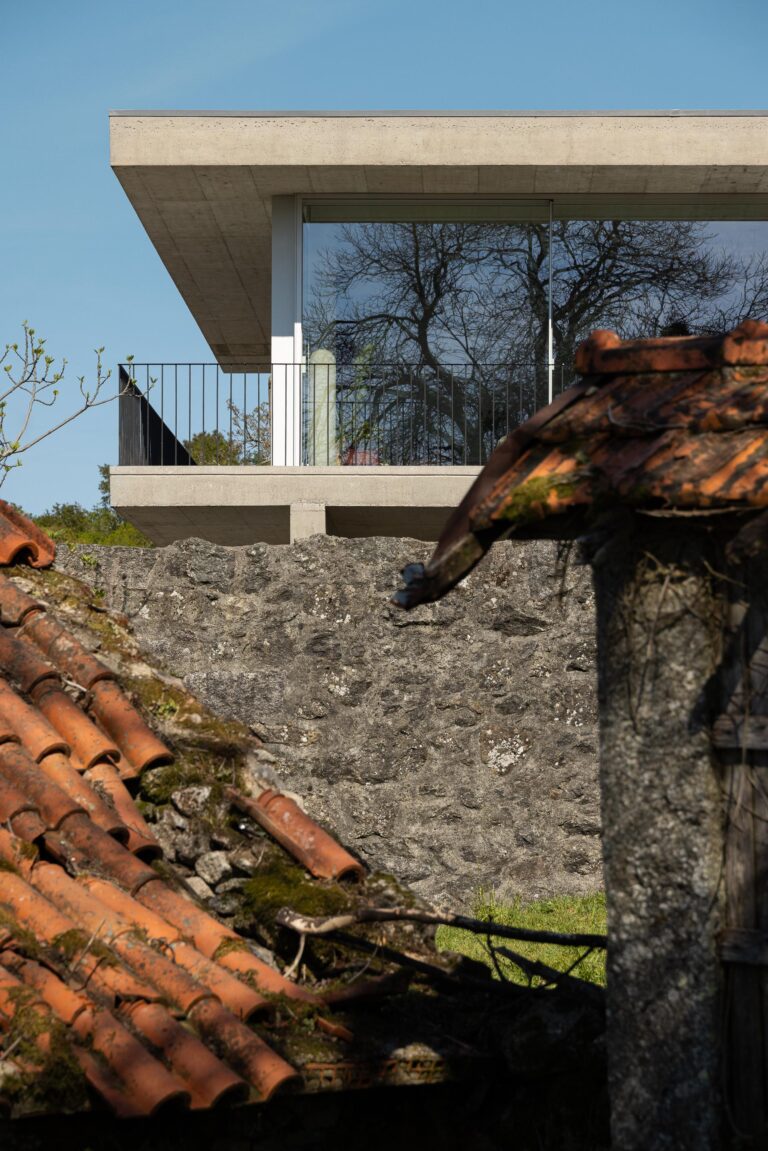 Detalhe da moradia com varanda em betão aparente e guarda-corpos metálicos, sobre muro de pedra tradicional e telhados antigos — CASA COQUEIRA arquitectura Tiago Sousa Arquitecto fotografia Ivo Tavares