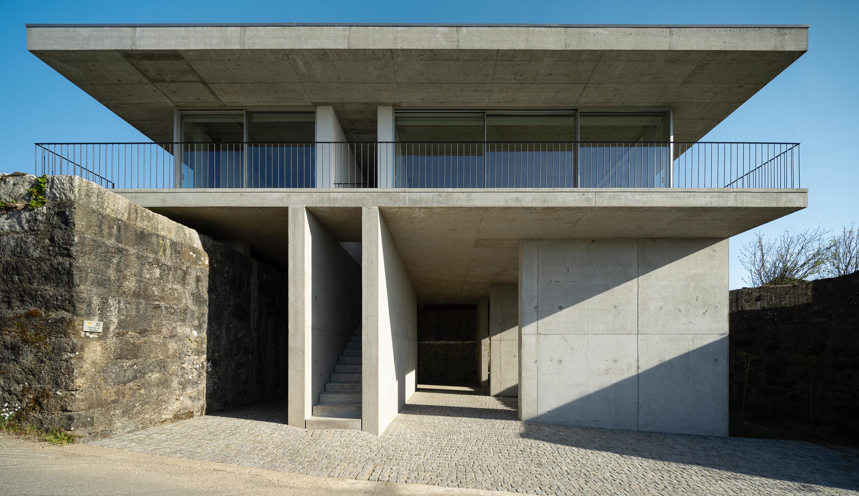 Fachada principal da moradia em betão aparente, com dois pisos, varanda superior e escadaria central. Muro de pedra lateral — CASA COQUEIRA arquitectura Tiago Sousa Arquitecto fotografia Ivo Tavares