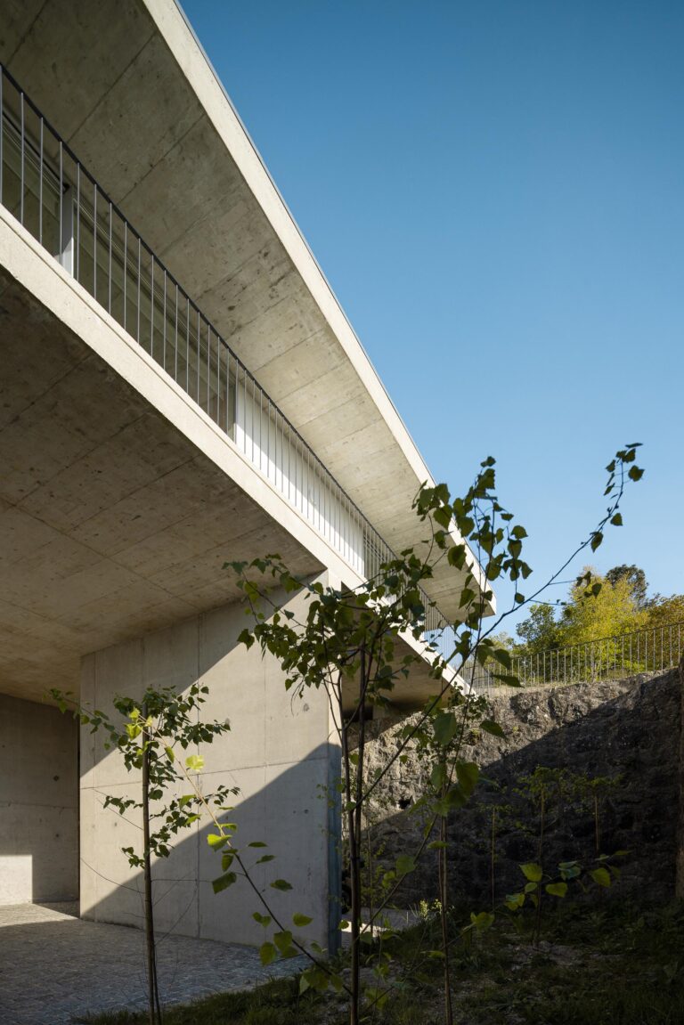 Perspetiva da moradia em betão aparente, com varanda em consola e muro de pedra, inserida na paisagem verde — CASA COQUEIRA arquitectura Tiago Sousa Arquitecto fotografia Ivo Tavares