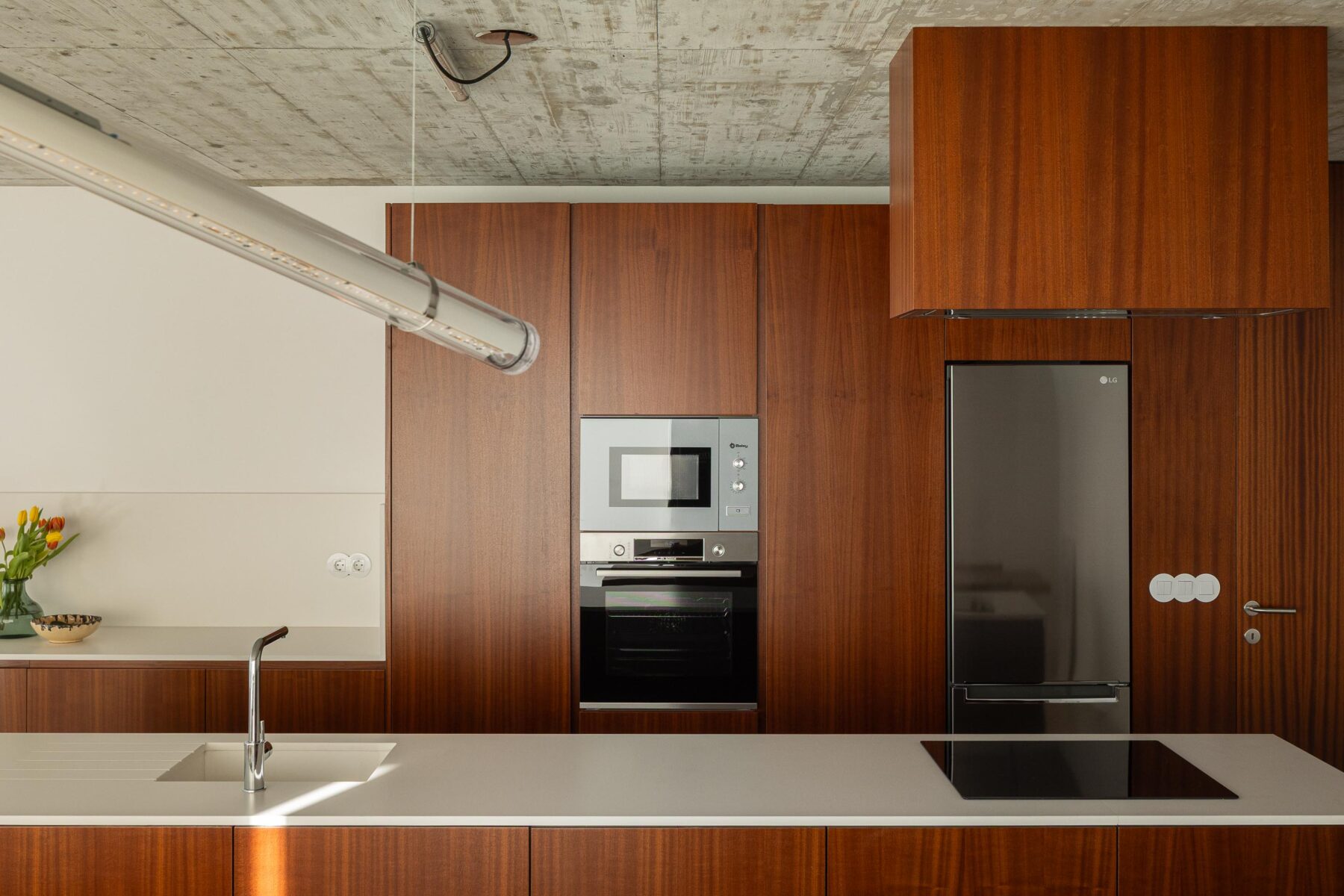 Detalhe da cozinha com armários em madeira escura, bancada branca, eletrodomésticos embutidos e teto em betão aparente — CASA COQUEIRA arquitectura Tiago Sousa Arquitecto fotografia Ivo Tavares
