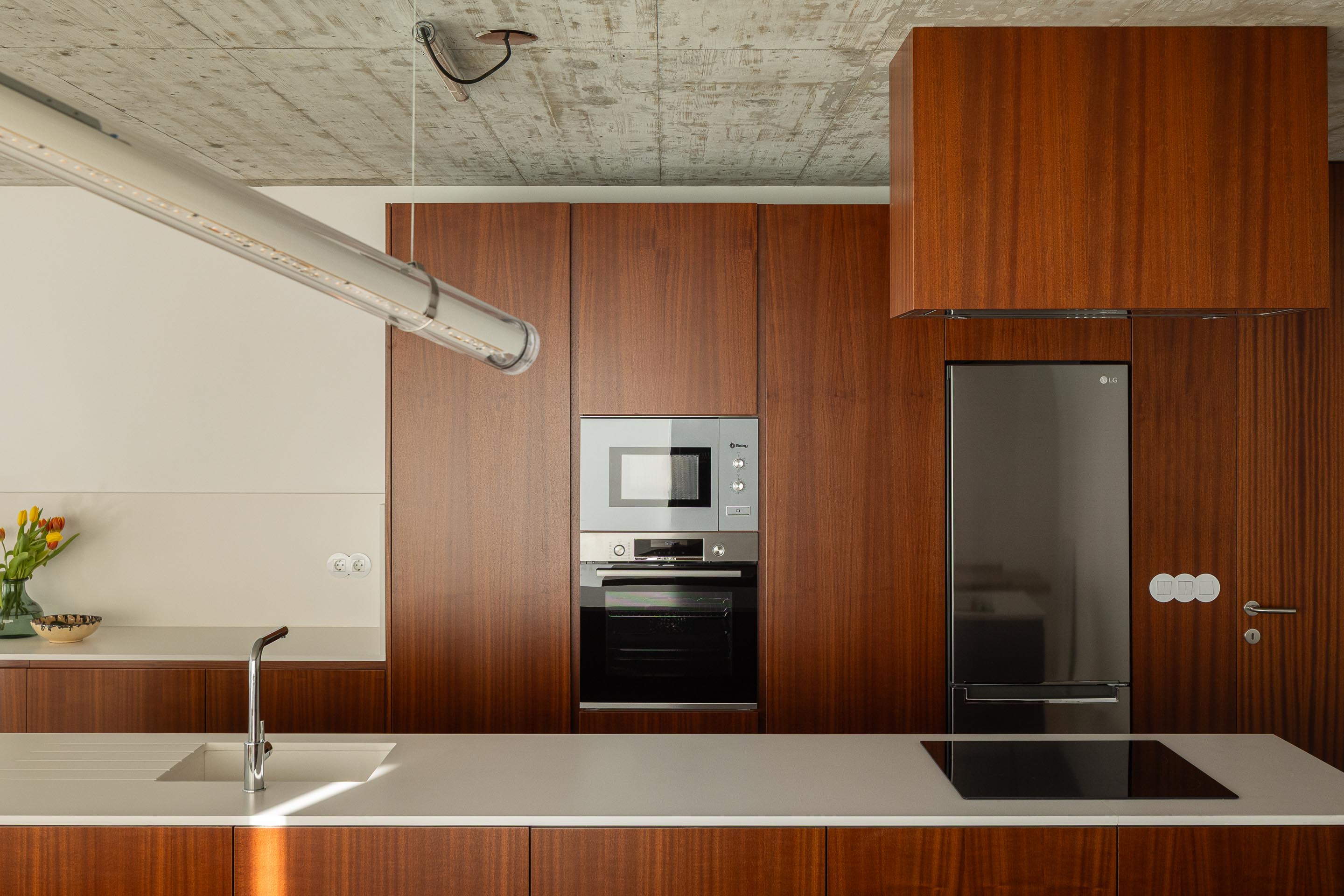Detalhe da cozinha com armários em madeira escura, bancada branca, eletrodomésticos embutidos e teto em betão aparente — CASA COQUEIRA arquitectura Tiago Sousa Arquitecto fotografia Ivo Tavares