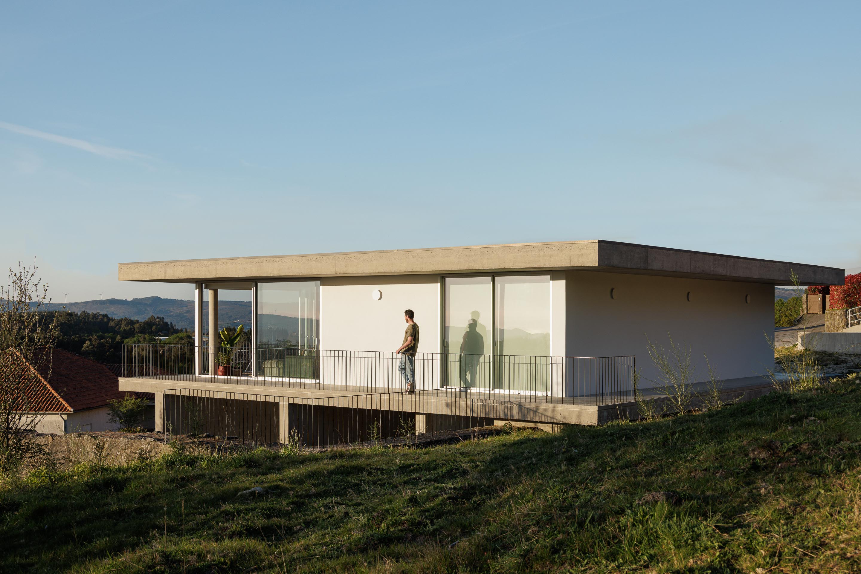 Moradia com fachada branca e vãos envidraçados, varanda em betão aparente e guarda-corpos metálicos. Homem na varanda — CASA COQUEIRA arquitectura Tiago Sousa Arquitecto fotografia Ivo Tavares