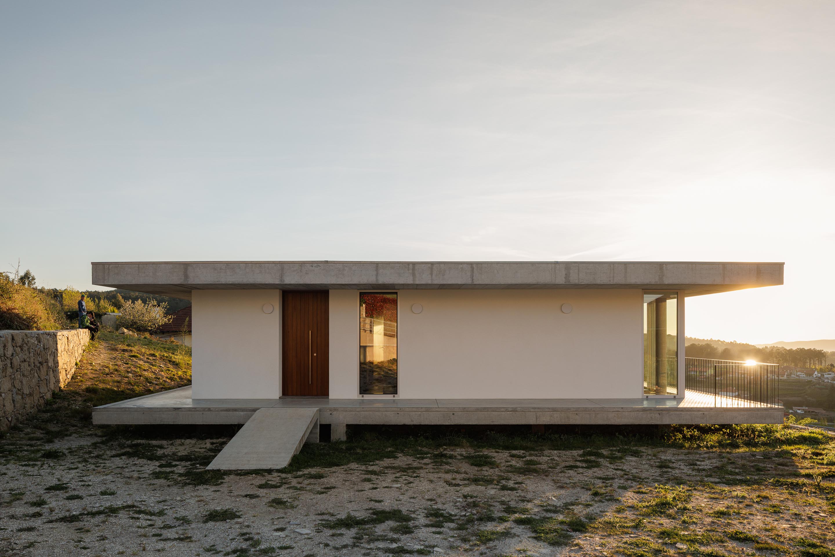 Fachada principal da moradia com paredes brancas, porta em madeira escura e vãos envidraçados, ao pôr do sol — CASA COQUEIRA arquitectura Tiago Sousa Arquitecto fotografia Ivo Tavares