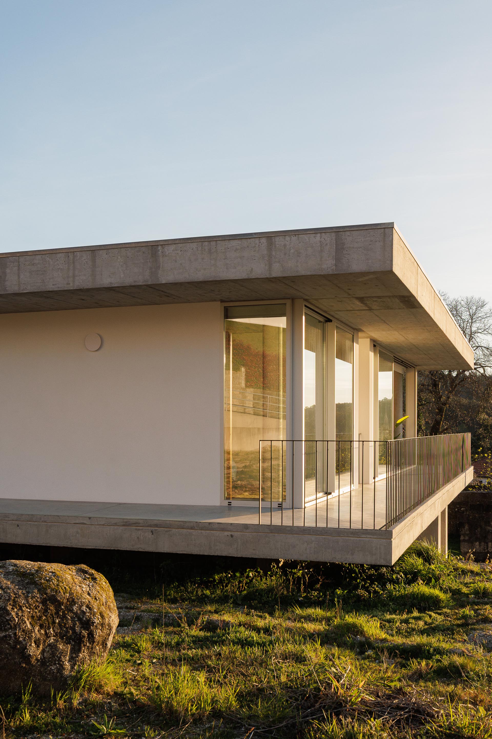 Canto da moradia com varanda em consola de betão aparente, grandes vãos envidraçados e luz dourada do pôr do sol — CASA COQUEIRA arquitectura Tiago Sousa Arquitecto fotografia Ivo Tavares