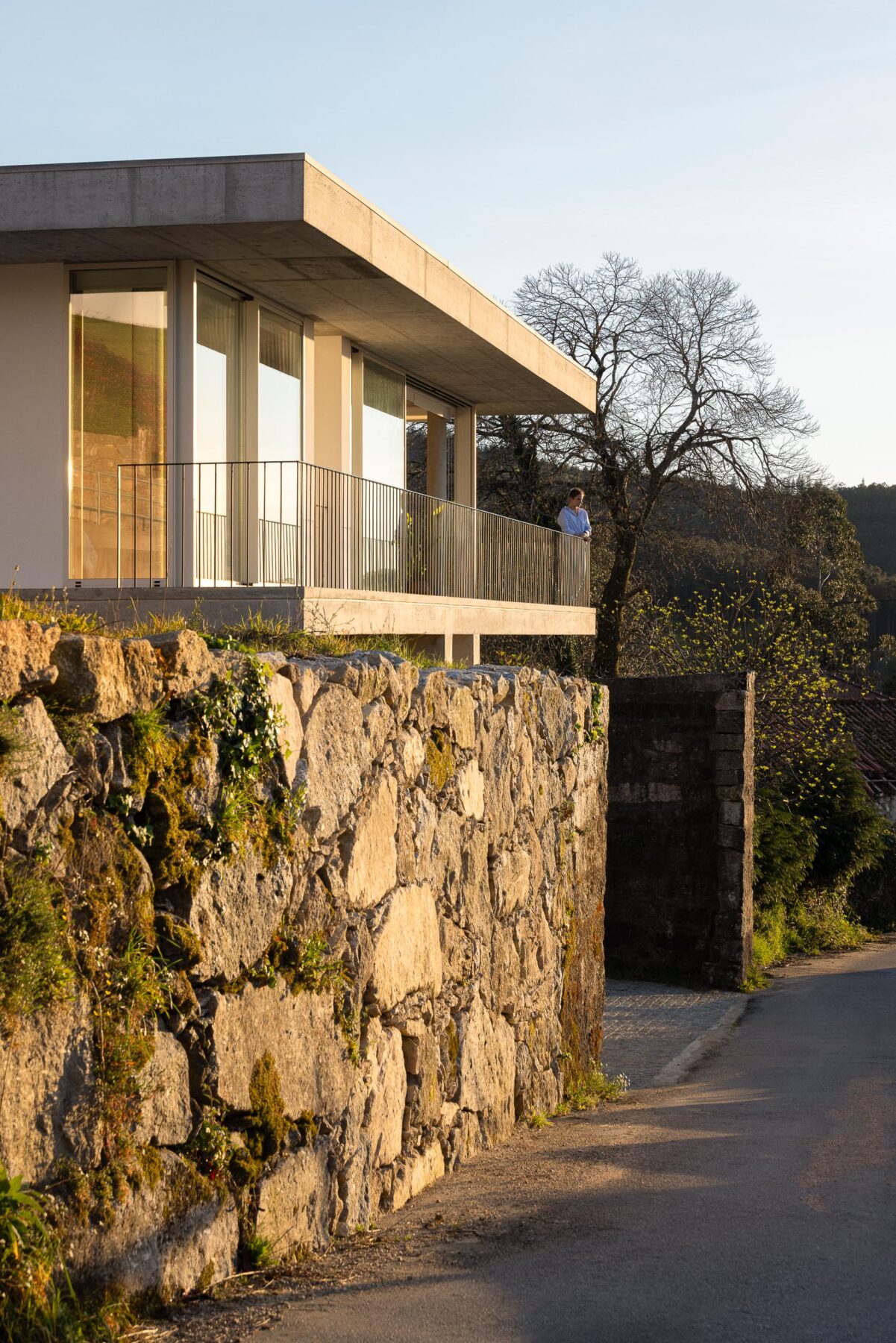 Moradia com varanda em betão aparente e guarda-corpos metálicos, sobre muro de pedra tradicional, ao pôr do sol — CASA COQUEIRA arquitectura Tiago Sousa Arquitecto fotografia Ivo Tavares