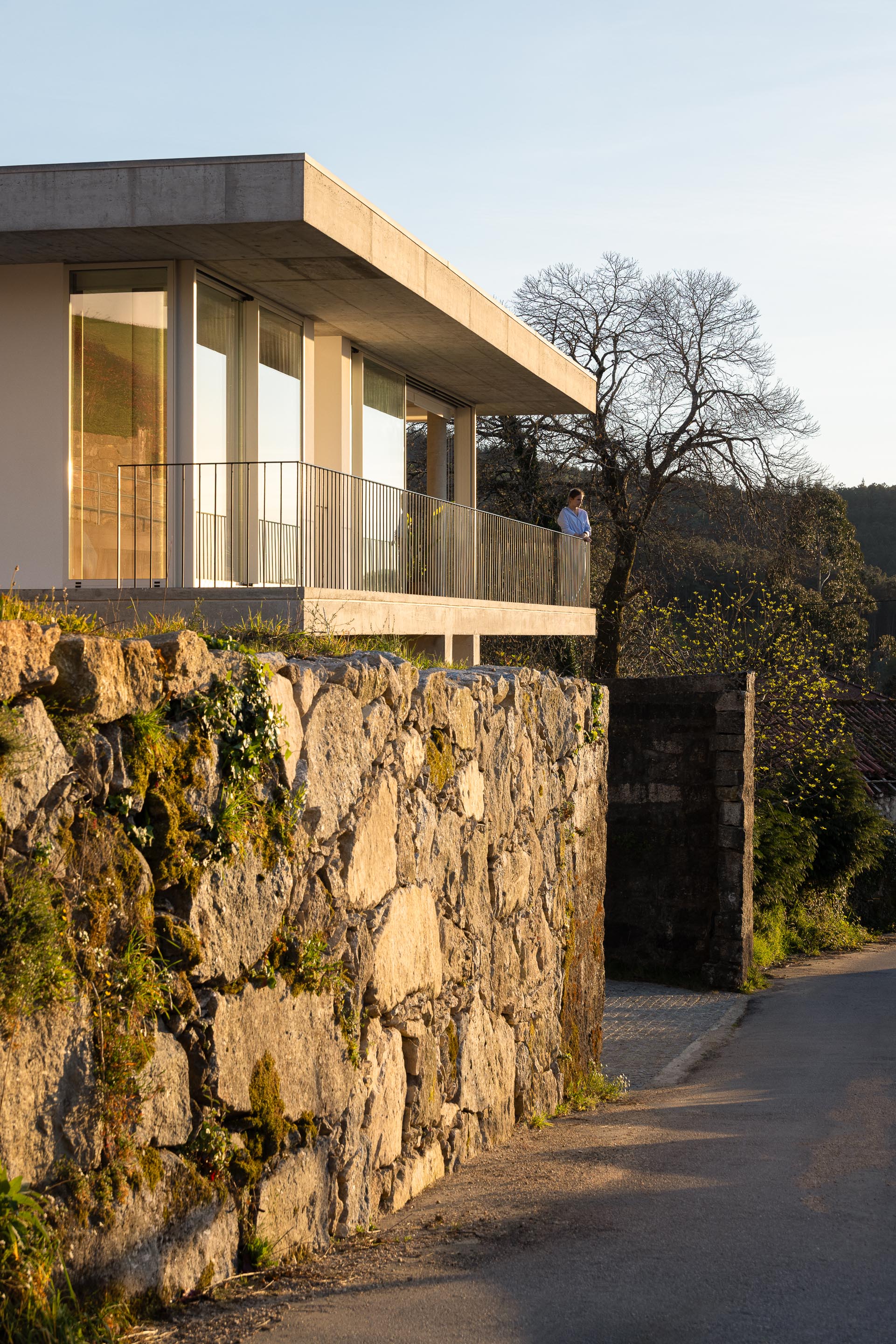 Moradia com varanda em betão aparente e guarda-corpos metálicos, sobre muro de pedra tradicional, ao pôr do sol — CASA COQUEIRA arquitectura Tiago Sousa Arquitecto fotografia Ivo Tavares