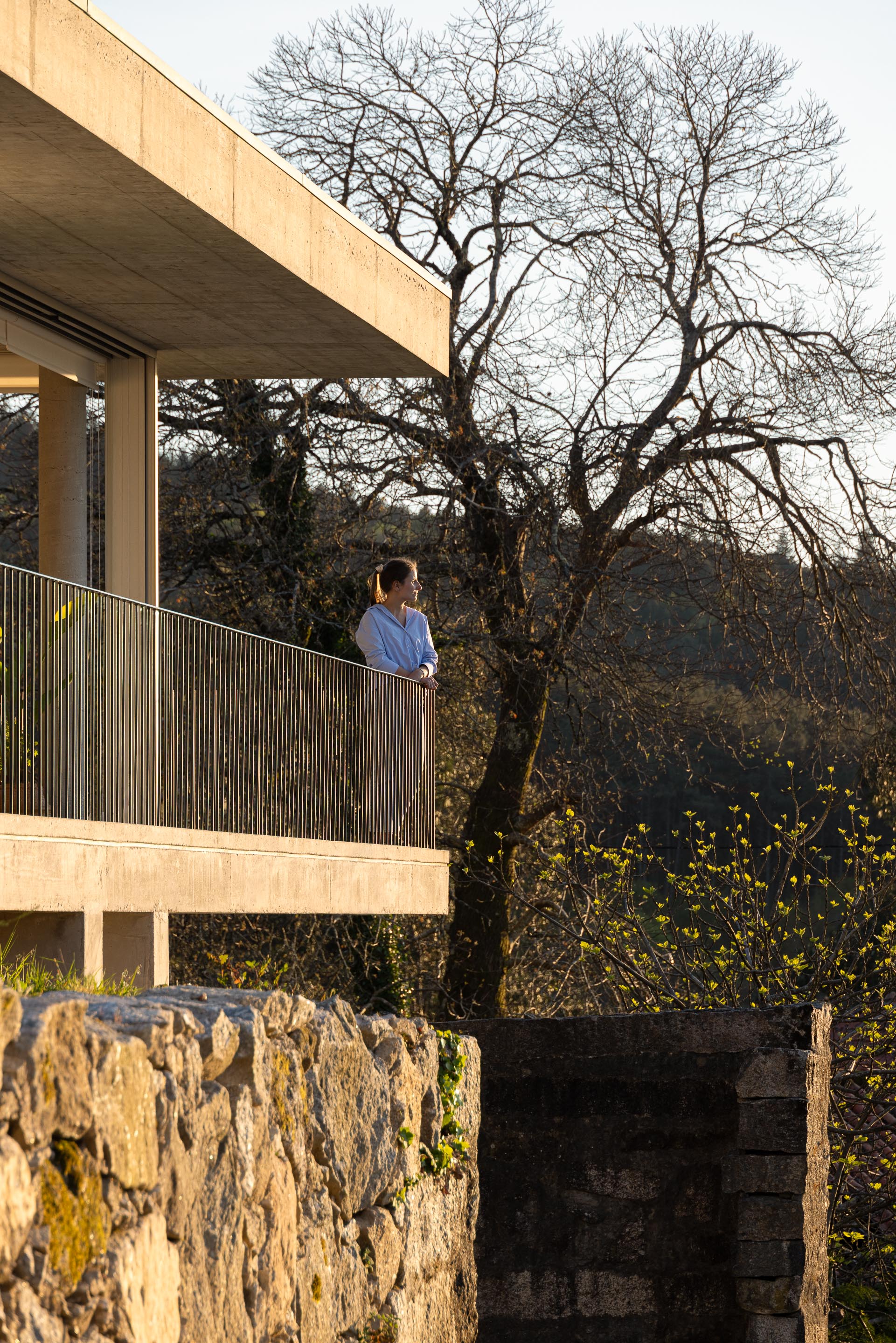 Mulher na varanda da moradia em betão aparente, com vista para a paisagem e árvore de folha caduca — CASA COQUEIRA arquitectura Tiago Sousa Arquitecto fotografia Ivo Tavares