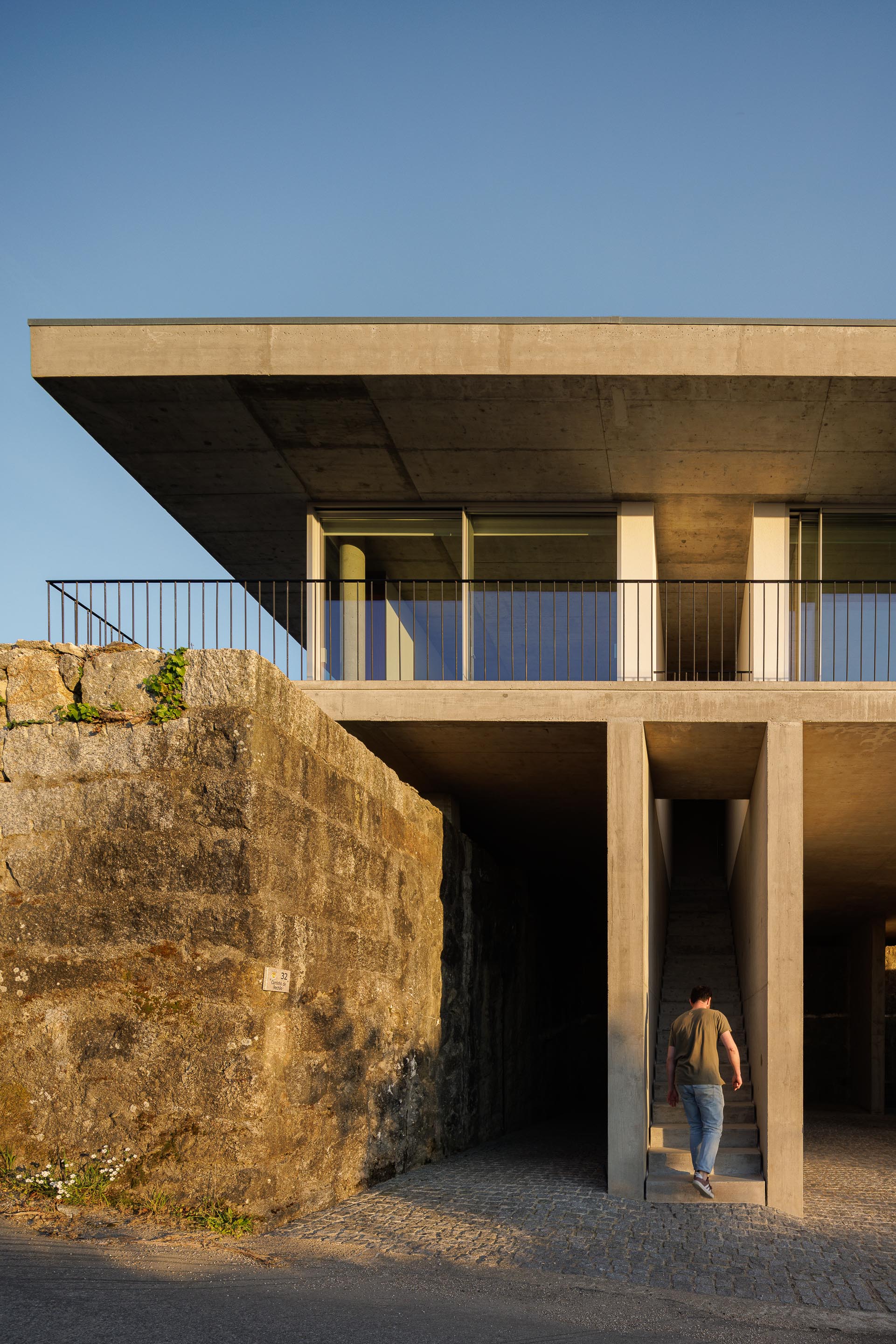 Homem a subir a escadaria de betão aparente, ladeada por muro de pedra, com a moradia elevada ao pôr do sol — CASA COQUEIRA arquitectura Tiago Sousa Arquitecto fotografia Ivo Tavares