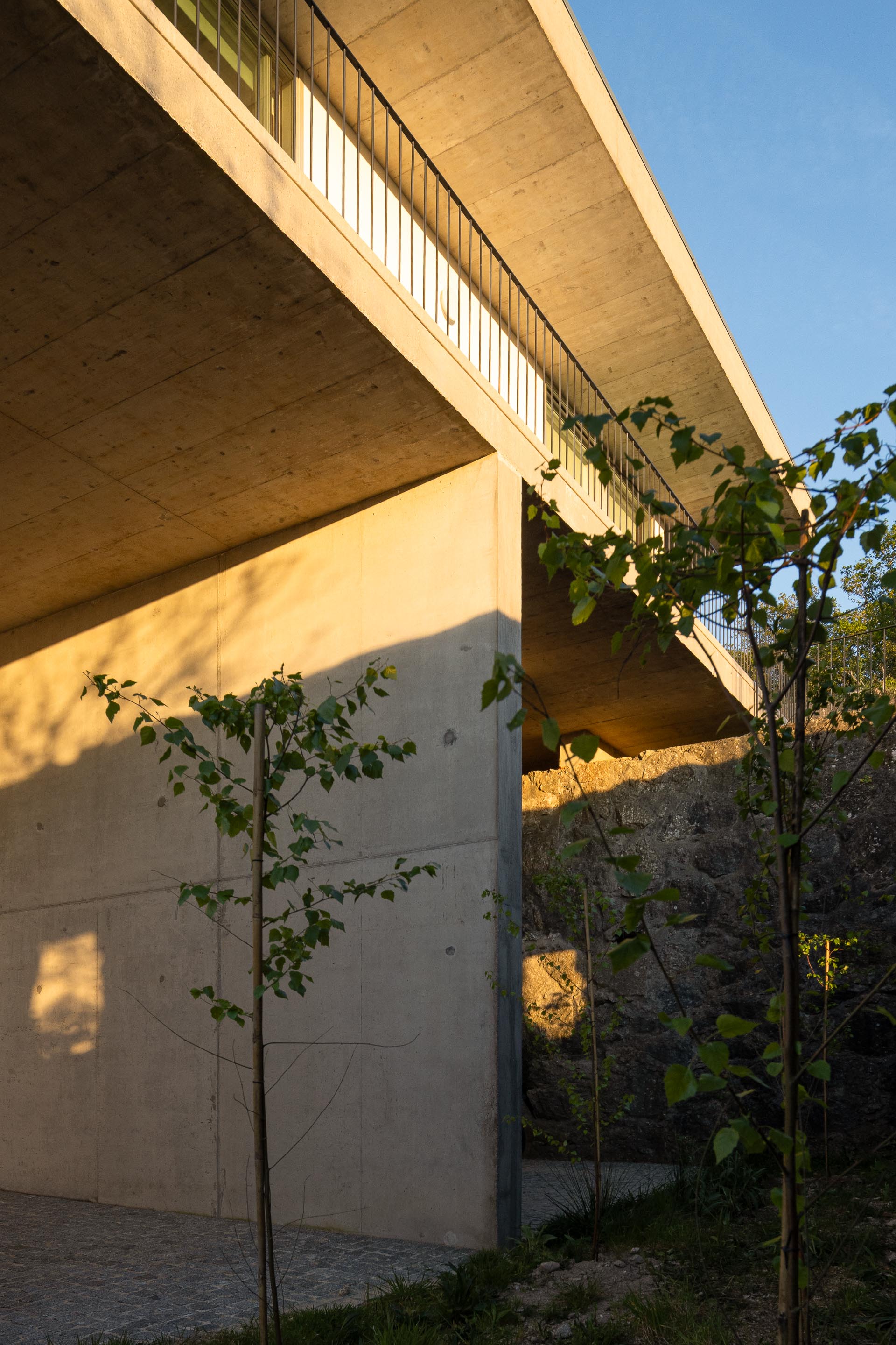 Moradia em betão aparente com varanda em consola, ladeada por muro de pedra e vegetação, sob luz dourada — CASA COQUEIRA arquitectura Tiago Sousa Arquitecto fotografia Ivo Tavares
