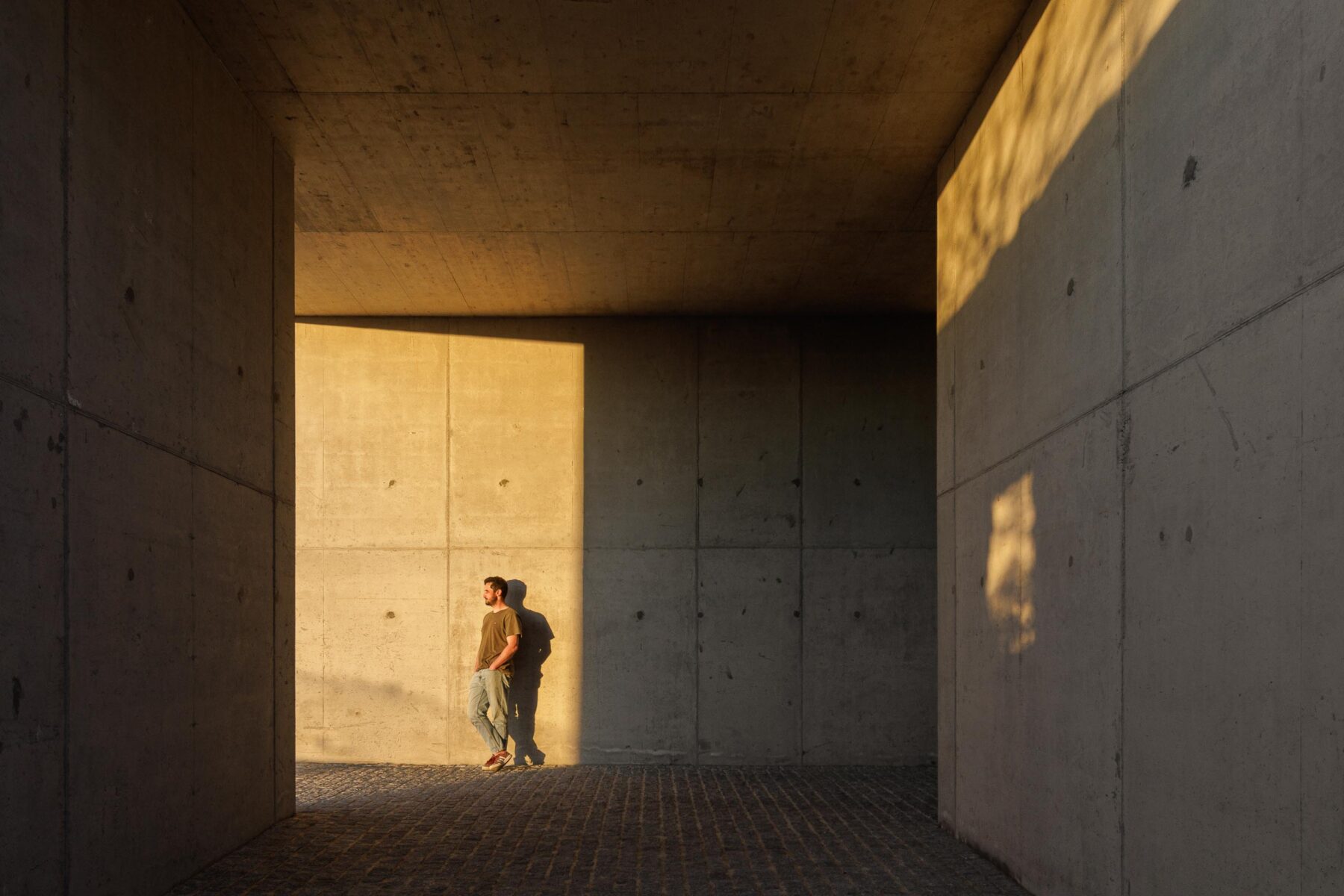Espaço coberto em betão aparente, com pavimento em calçada, banhado pela luz dourada do pôr do sol e sombras longas — CASA COQUEIRA arquitectura Tiago Sousa Arquitecto fotografia Ivo Tavares