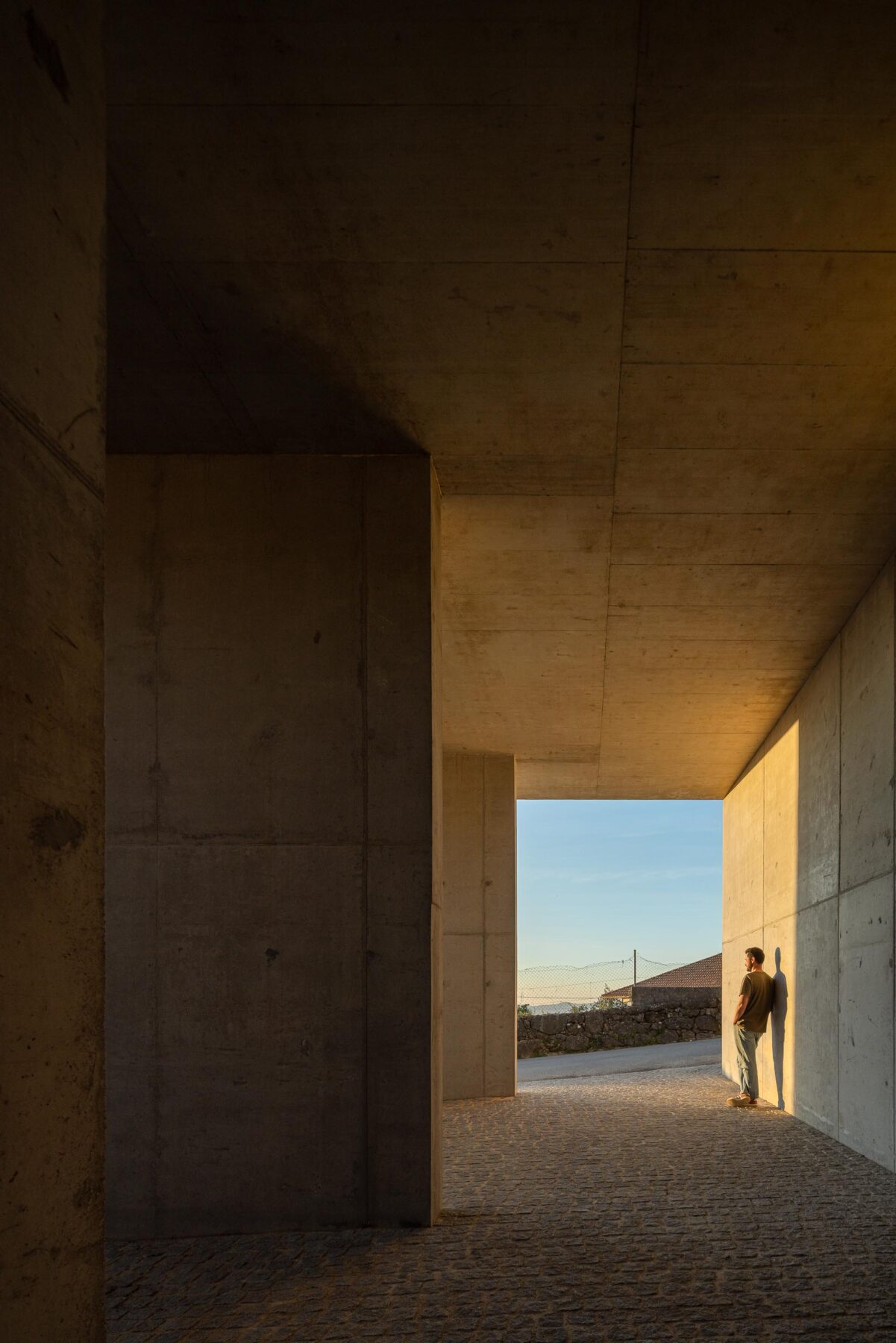 Espaço coberto em betão aparente, com pilares e pavimento em calçada, banhado pela luz do pôr do sol — CASA COQUEIRA arquitectura Tiago Sousa Arquitecto fotografia Ivo Tavares