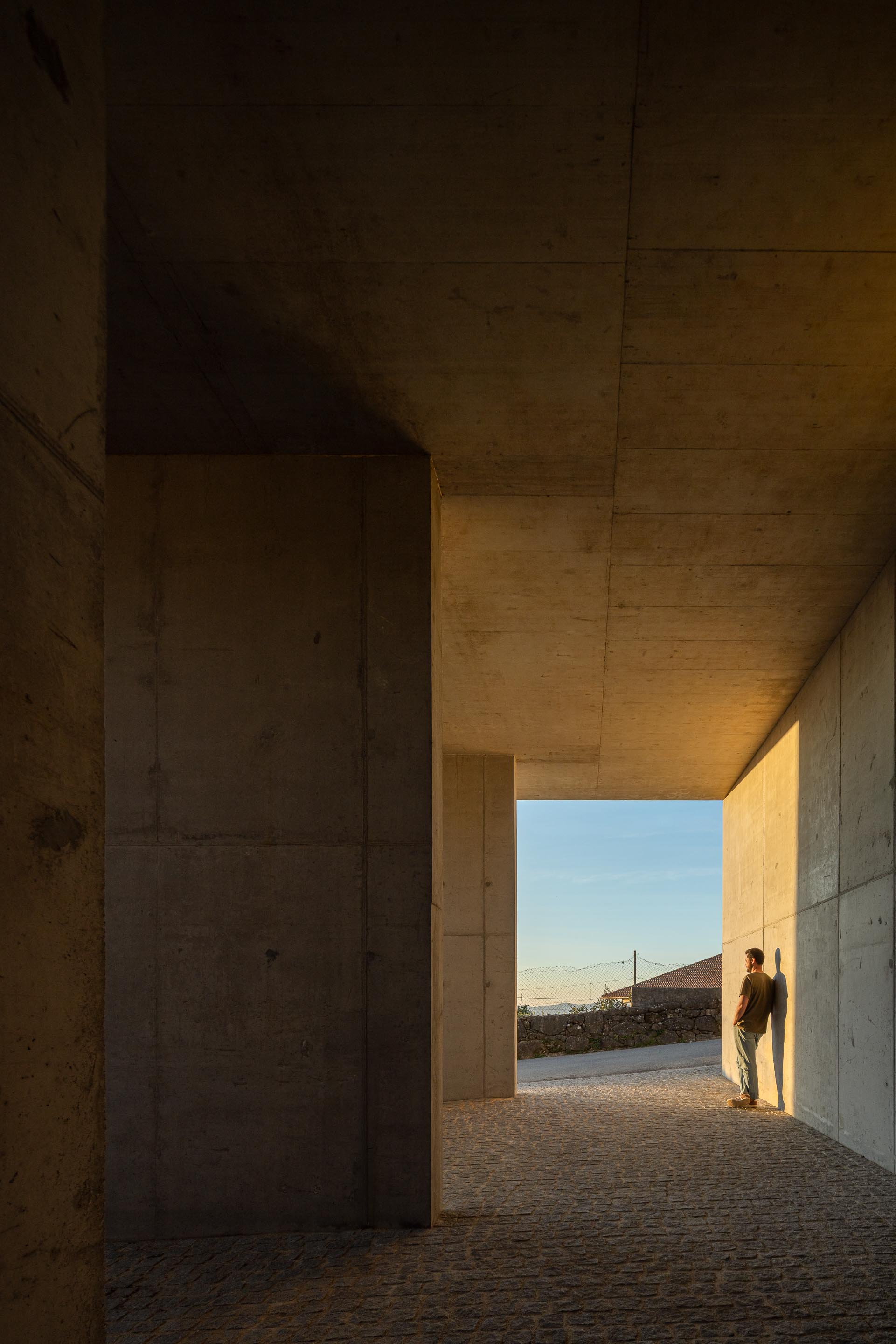 Espaço coberto em betão aparente, com pilares e pavimento em calçada, banhado pela luz do pôr do sol — CASA COQUEIRA arquitectura Tiago Sousa Arquitecto fotografia Ivo Tavares