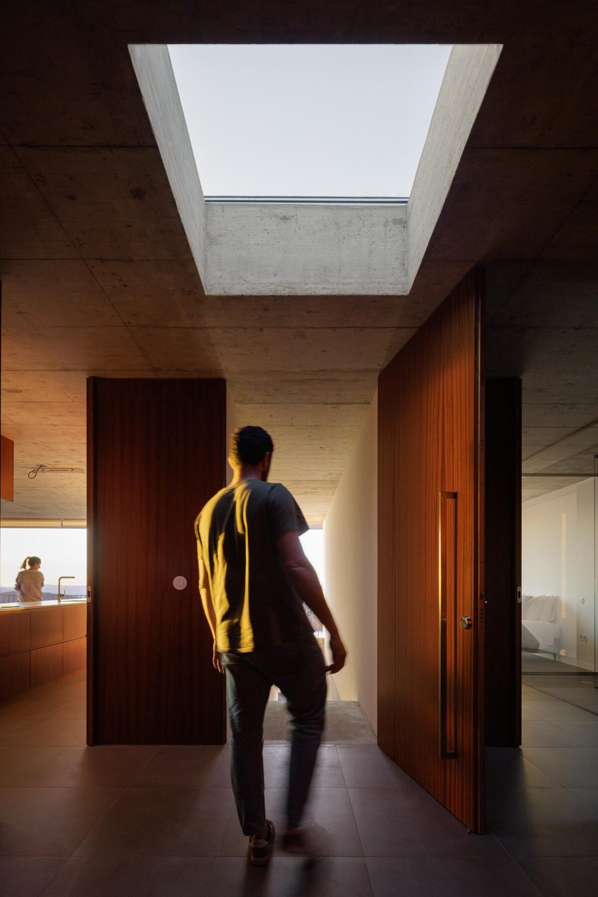 Interior da moradia com teto em betão aparente e claraboia, portas em madeira escura. Homem a caminhar — CASA COQUEIRA arquitectura Tiago Sousa Arquitecto fotografia Ivo Tavares
