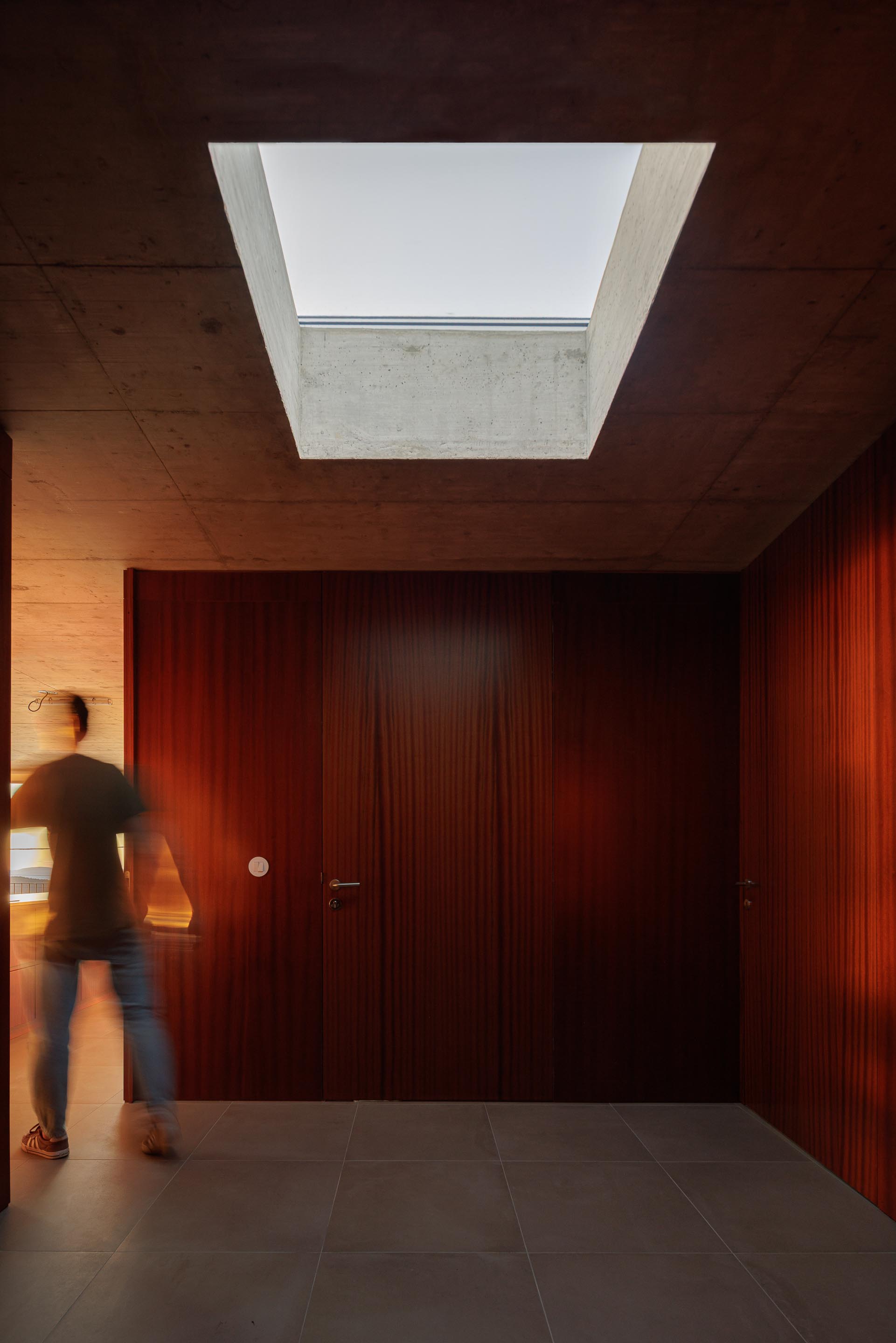 Interior da moradia com teto em betão aparente e claraboia, portas em madeira escura. Homem em movimento — CASA COQUEIRA arquitectura Tiago Sousa Arquitecto fotografia Ivo Tavares