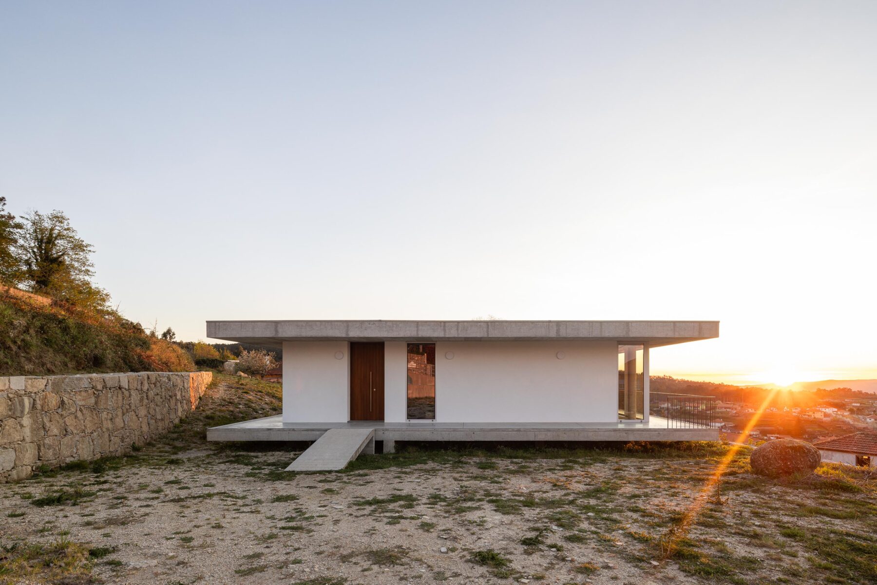 Fachada principal da moradia com paredes brancas, porta em madeira escura e vãos envidraçados, sob luz do pôr do sol — CASA COQUEIRA arquitectura Tiago Sousa Arquitecto fotografia Ivo Tavares