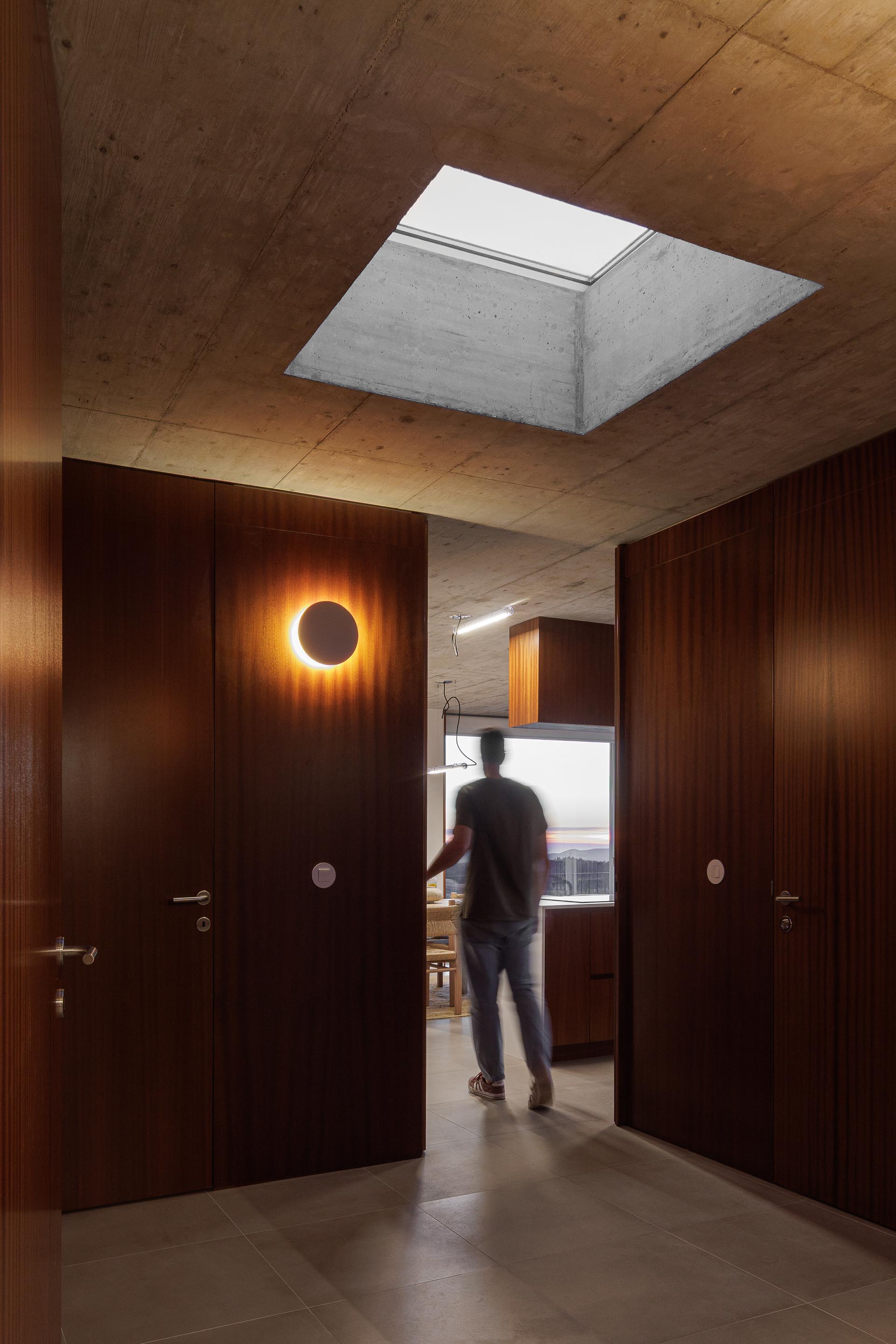 Corredor com teto em betão aparente e claraboia, portas em madeira escura e candeeiro de parede circular — CASA COQUEIRA arquitectura Tiago Sousa Arquitecto fotografia Ivo Tavares