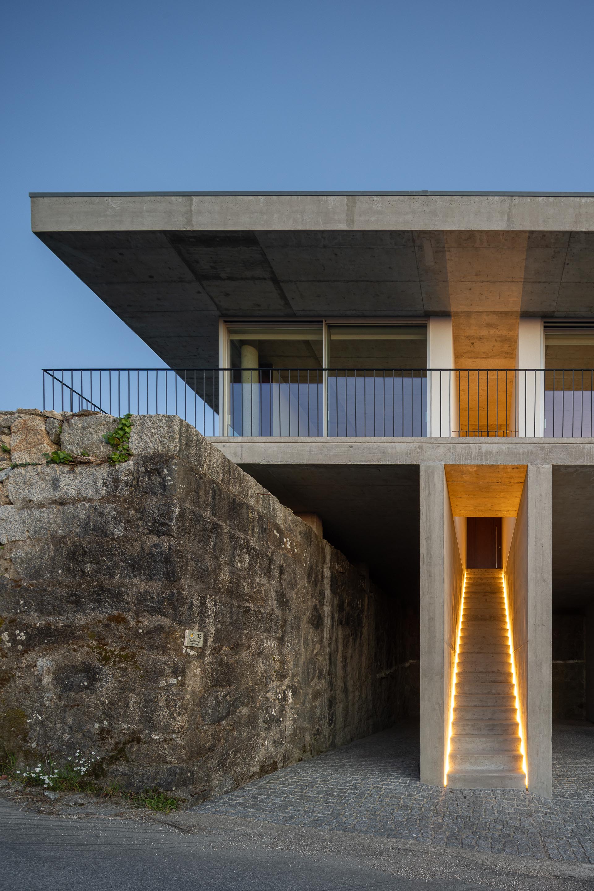 Escadaria exterior de betão aparente, iluminada por luz linear, sob a moradia elevada, ao anoitecer — CASA COQUEIRA arquitectura Tiago Sousa Arquitecto fotografia Ivo Tavares