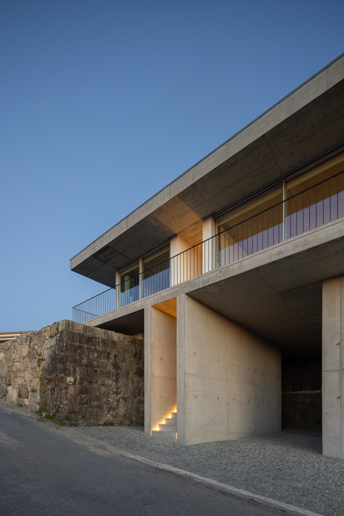 Moradia em betão aparente com varanda e escadaria iluminada, ladeada por muro de pedra, ao anoitecer — CASA COQUEIRA arquitectura Tiago Sousa Arquitecto fotografia Ivo Tavares