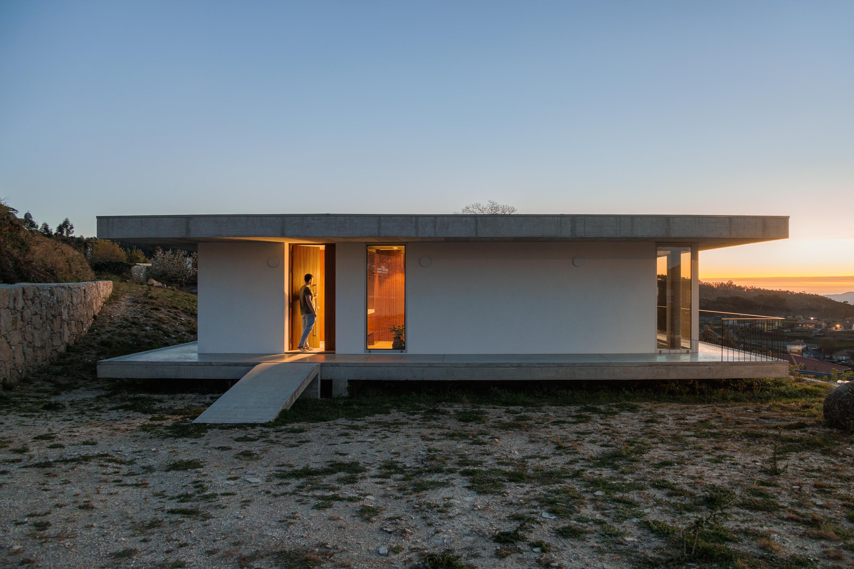 Fachada principal da moradia com paredes brancas, porta em madeira escura e vãos envidraçados, iluminada ao anoitecer — CASA COQUEIRA arquitectura Tiago Sousa Arquitecto fotografia Ivo Tavares