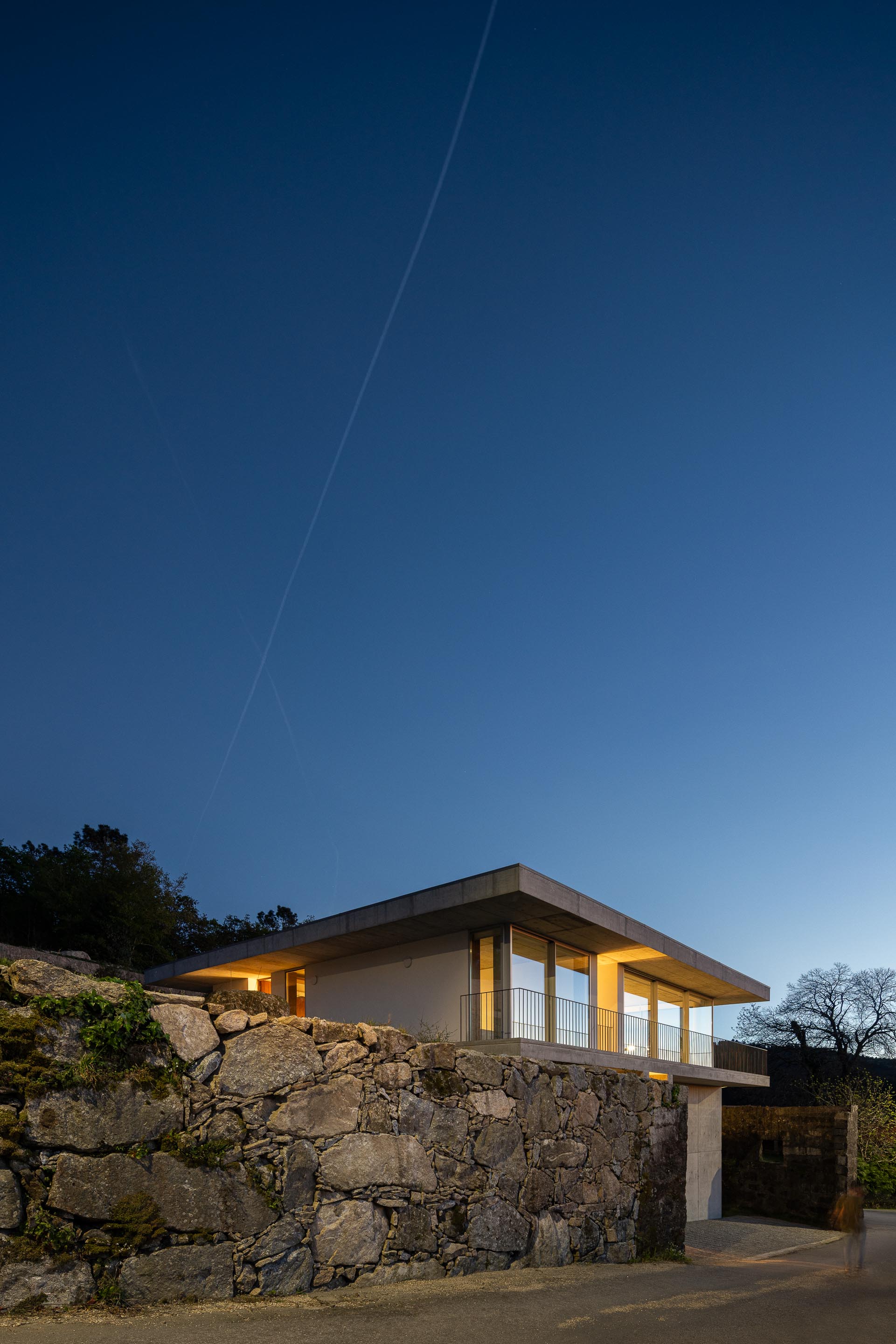 Moradia em betão aparente e paredes brancas, com varanda e vãos envidraçados, iluminada ao anoitecer, sobre muro de pedra — CASA COQUEIRA arquitectura Tiago Sousa Arquitecto fotografia Ivo Tavares
