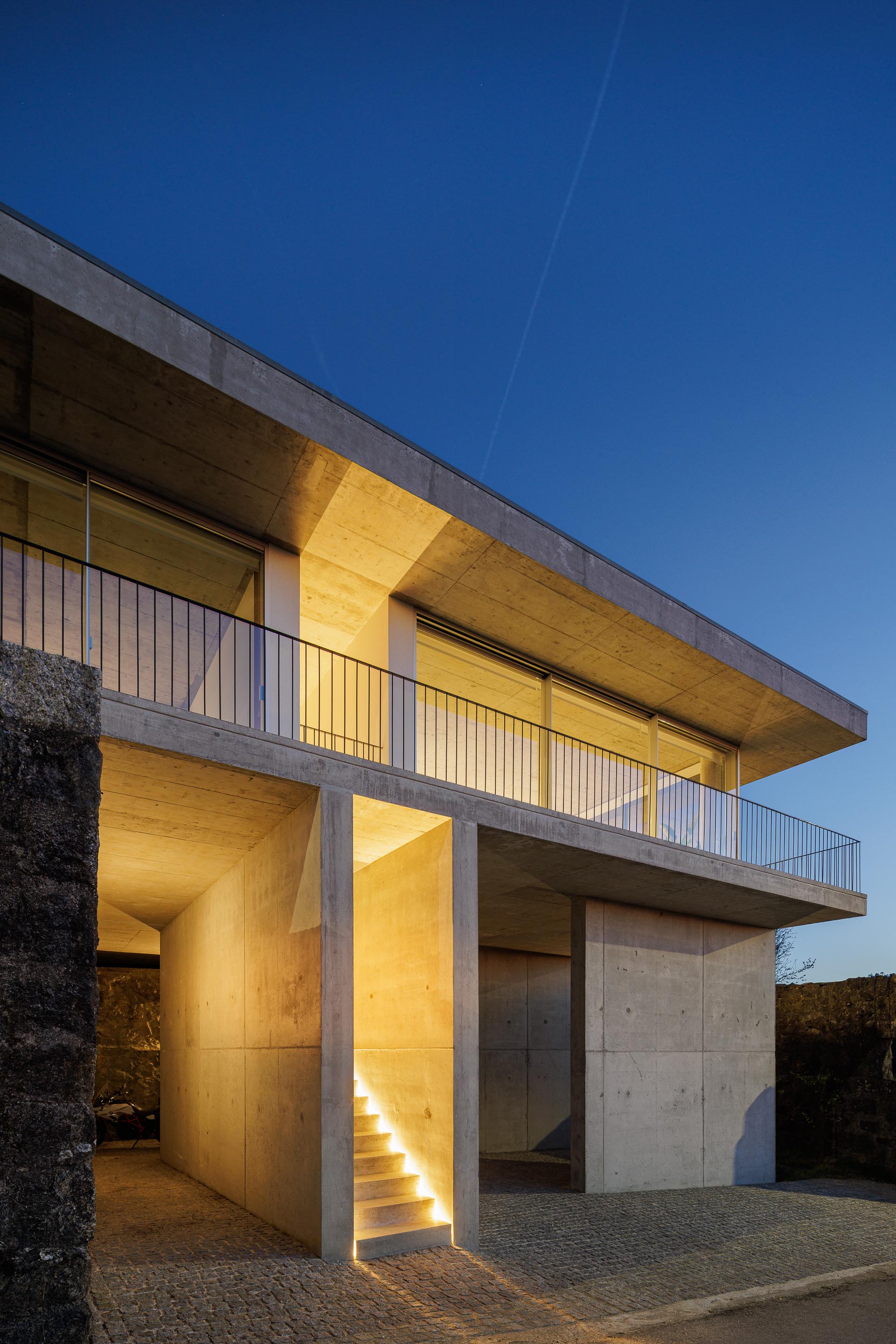 Moradia em betão aparente com varanda e vãos envidraçados, iluminada, sobre muro de pedra, ao anoitecer — CASA COQUEIRA arquitectura Tiago Sousa Arquitecto fotografia Ivo Tavares