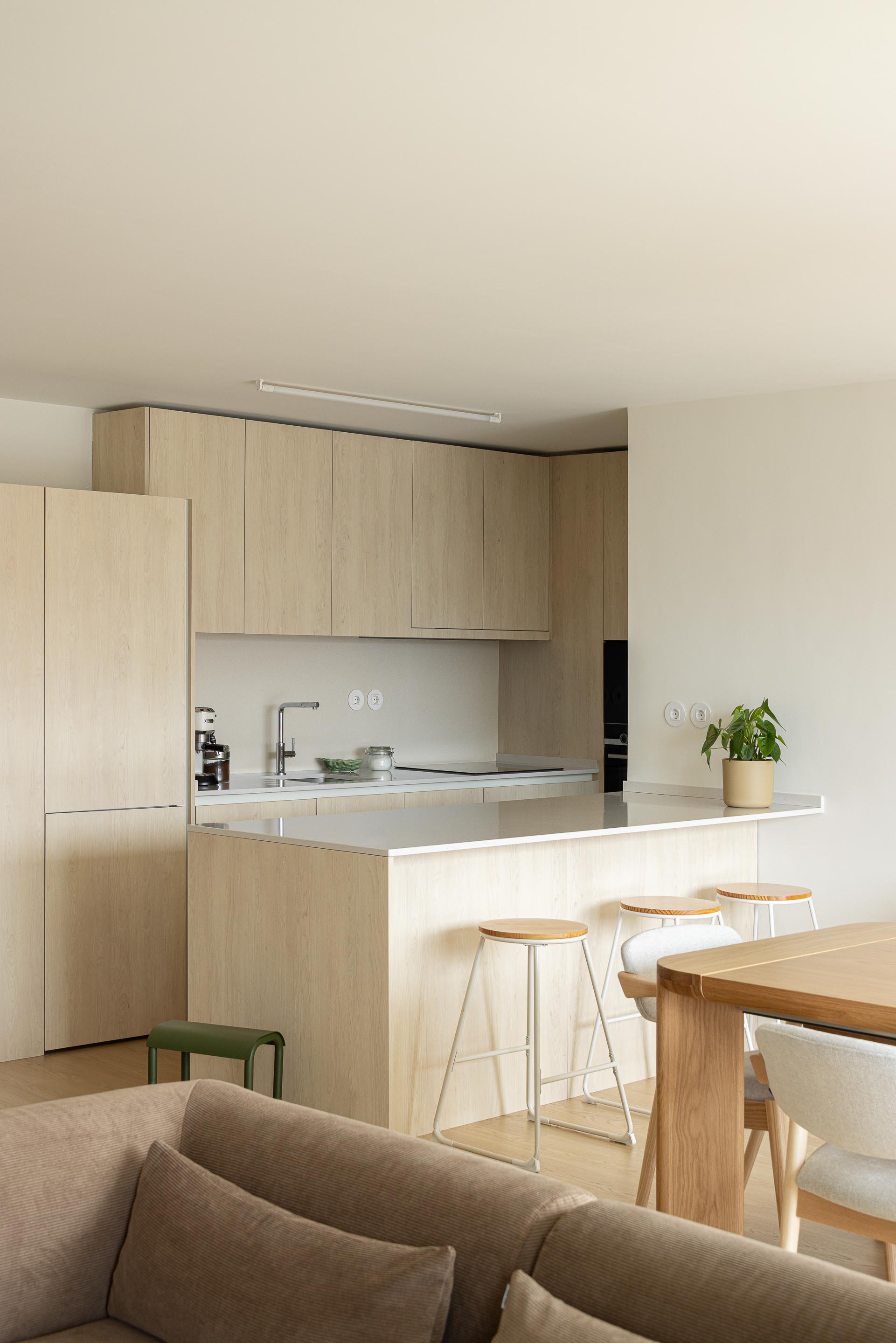 Cozinha funcional com armários em madeira clara e ilha central com bancada branca, aberta para a área de refeições — Costa Cabral Building arquitectura ENTRETEMPOS + ATA Atelier fotografia Ivo Tavares