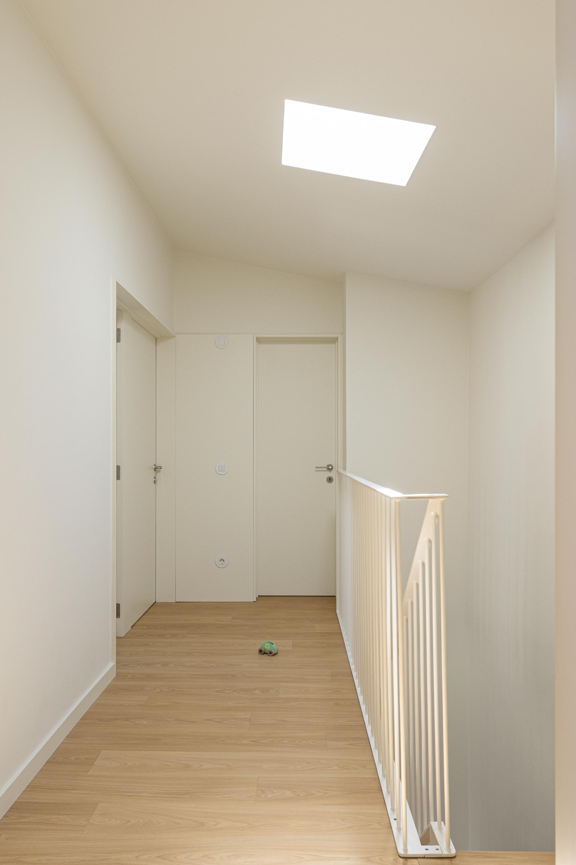 Corredor iluminado por vão zenital, com pavimento em madeira clara e gradil metálico branco da escada — Costa Cabral Building arquitectura ENTRETEMPOS + ATA Atelier fotografia Ivo Tavares