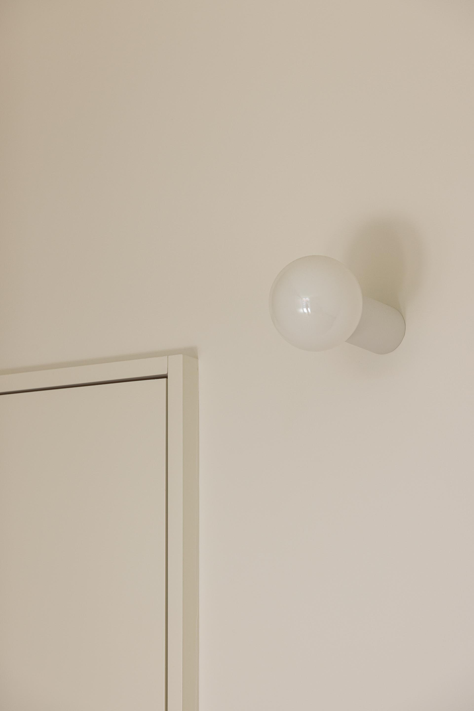 Detalhe de candeeiro de parede esférico branco sobre parede de cor clara, junto a um vão de porta — Costa Cabral Building arquitectura ENTRETEMPOS + ATA Atelier fotografia Ivo Tavares