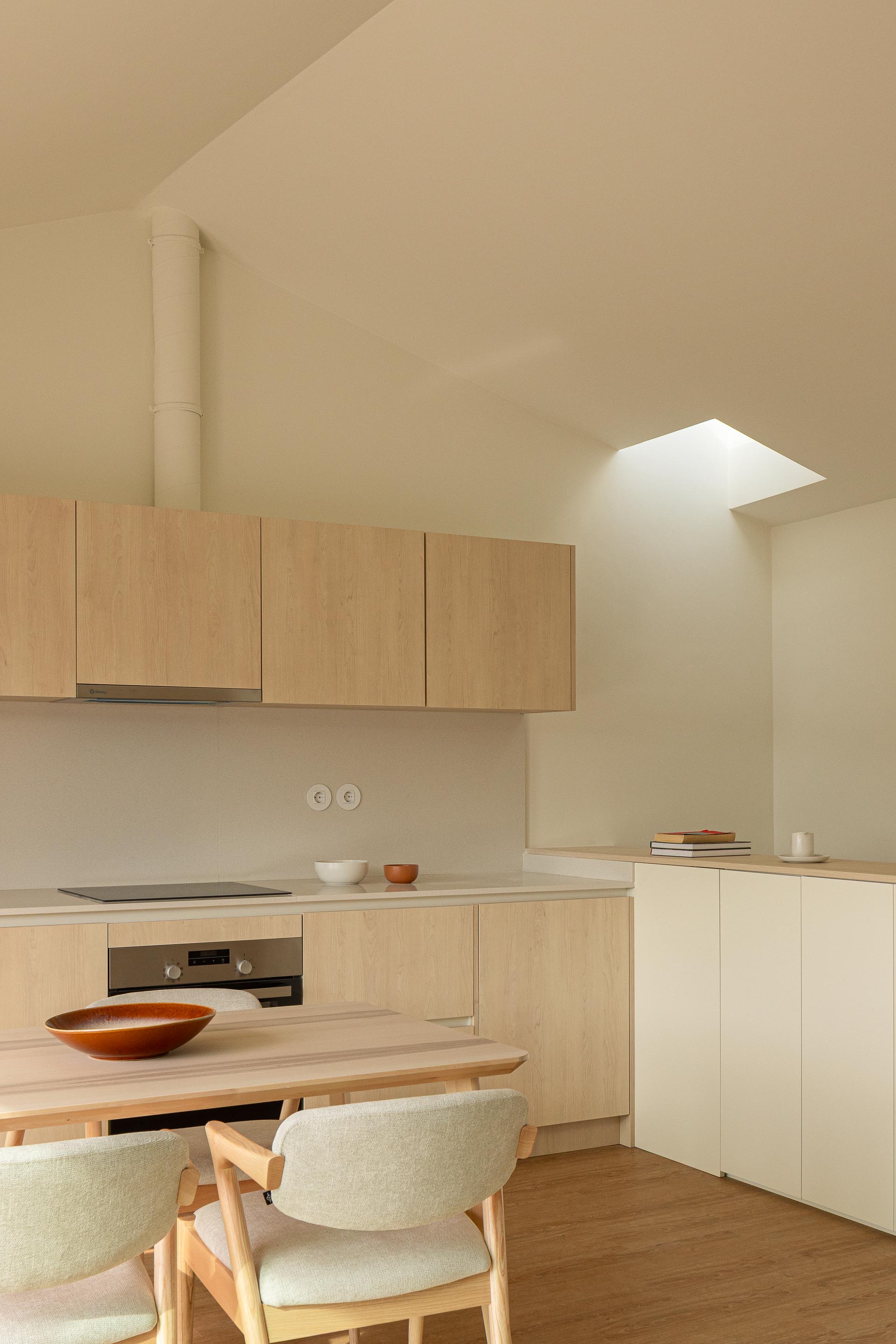 Cozinha no pavimento superior com armários em madeira clara, bancada branca e iluminação zenital através de vão na cobertura inclinada — Costa Cabral Building arquitectura ENTRETEMPOS + ATA Atelier fotografia Ivo Tavares