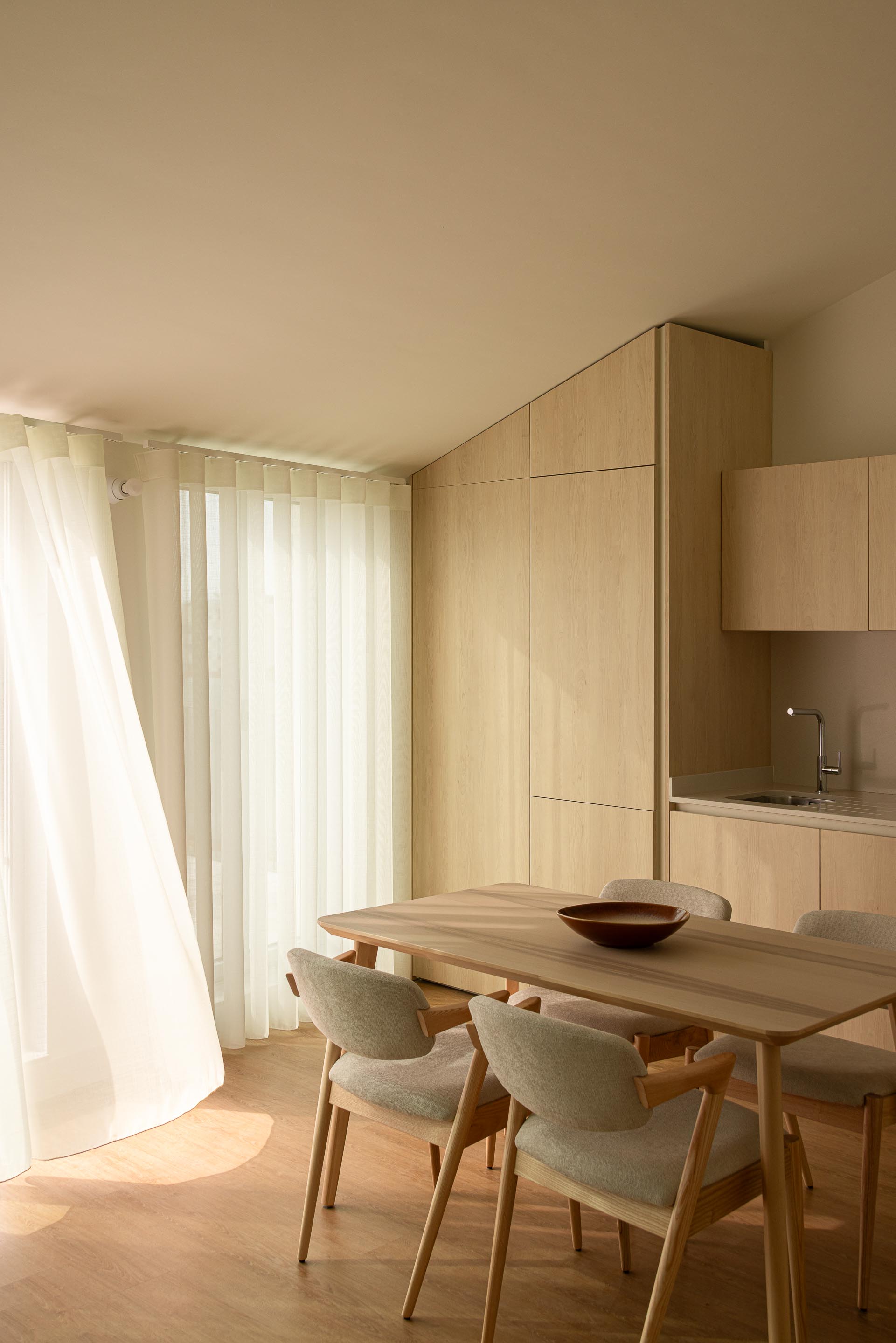 Cozinha e sala de jantar no pavimento superior, com armários em madeira clara e cortinas brancas a filtrar a luz natural — Costa Cabral Building arquitectura ENTRETEMPOS + ATA Atelier fotografia Ivo Tavares