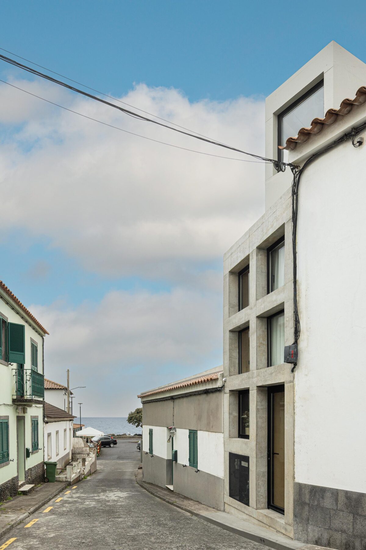 Fachada em betão aparente cinzento com caixilharia preta, inserida em rua inclinada com vista para o mar — MINI ATALHADA arquitectura SO Arquitetura & Design fotografia Ivo Tavares