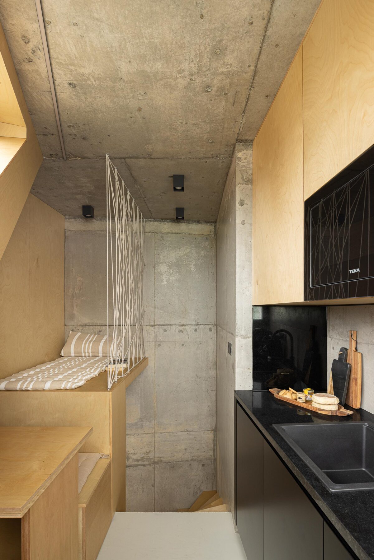 Cozinha com bancada preta e armários em madeira clara, adjacente a zona de estar em betão aparente — MINI ATALHADA arquitectura SO Arquitetura & Design fotografia Ivo Tavares