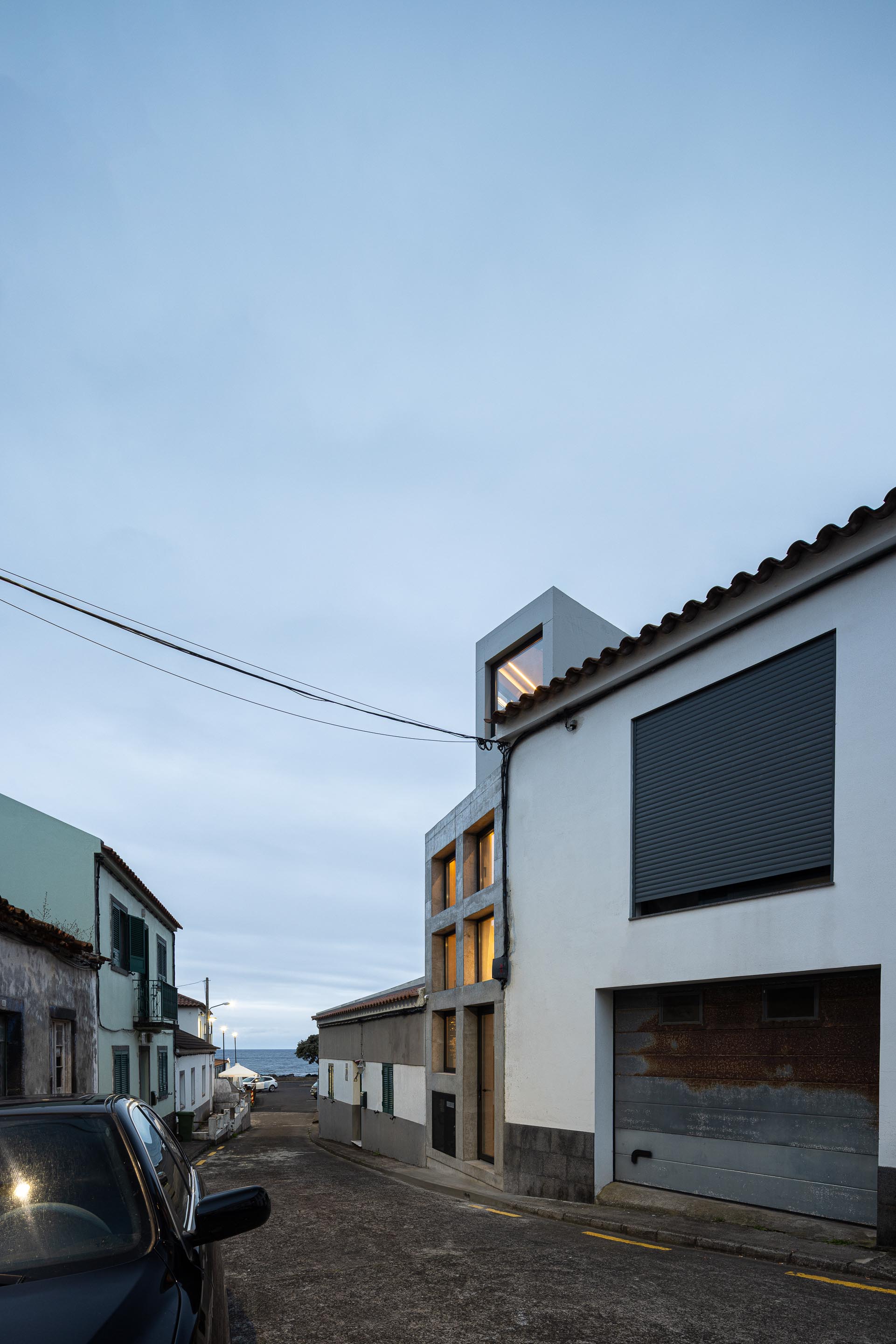Fachada em betão aparente cinzento à noite, inserida em rua inclinada com vista para o mar — MINI ATALHADA arquitectura SO Arquitetura & Design fotografia Ivo Tavares
