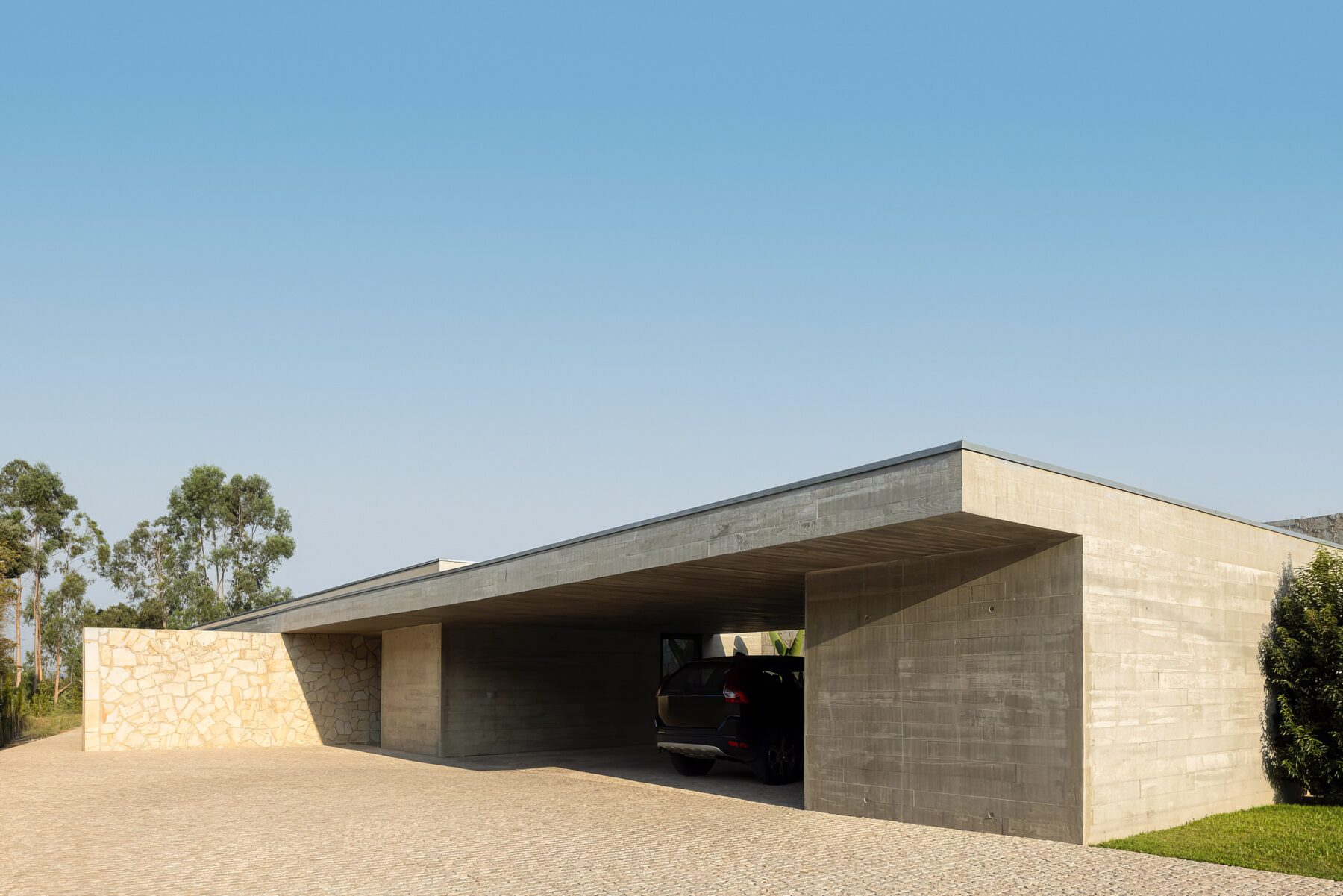 Entrada da moradia com alpendre em betão aparente, pavimento em calçada e paredes em alvenaria de granito — Quinta da Casa (Habitação QC)