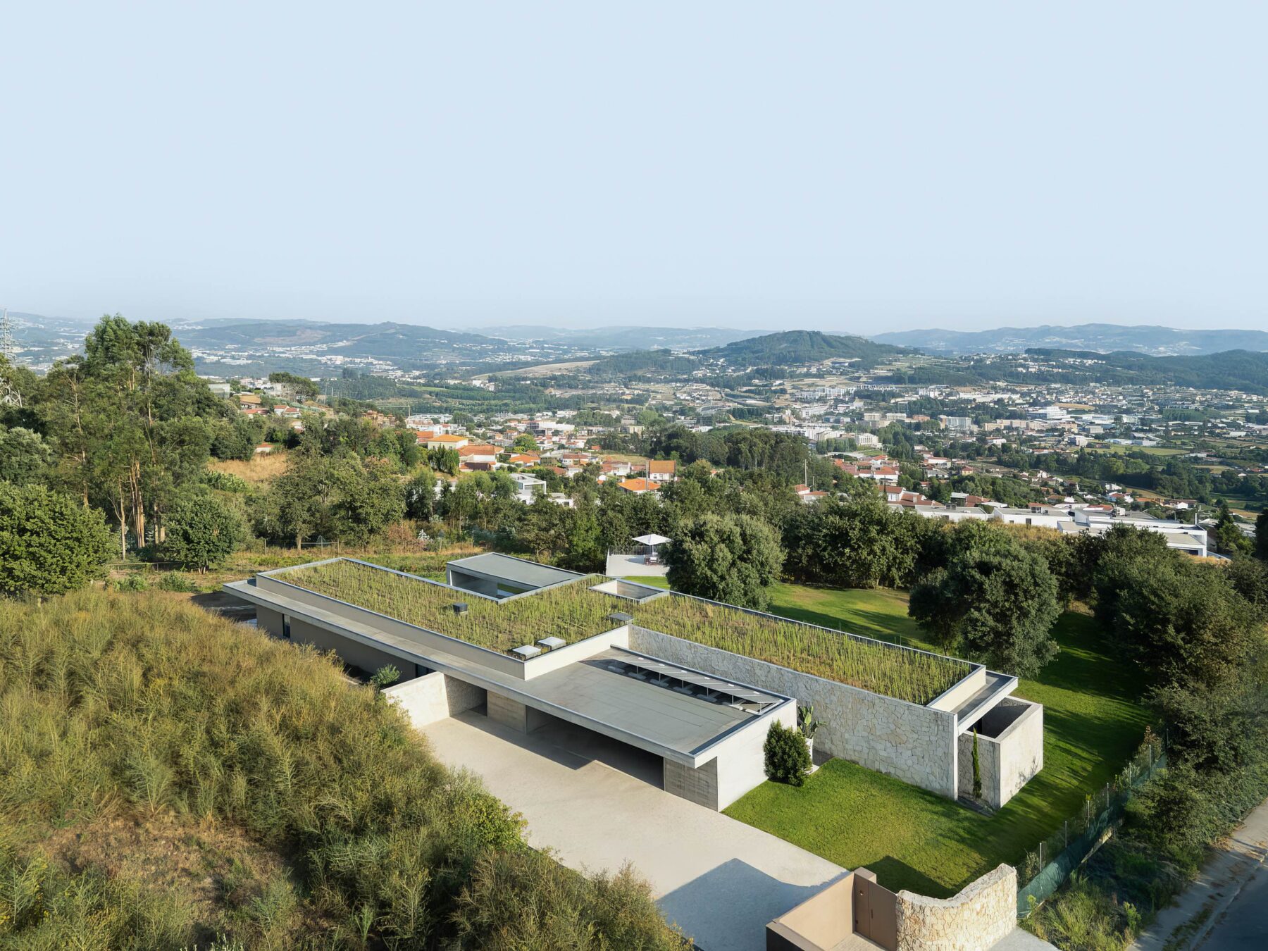 Vista aérea da moradia com cobertura ajardinada, volumes em betão aparente e alvenaria de granito, integrada na paisagem — Quinta da Casa (Habitação QC)