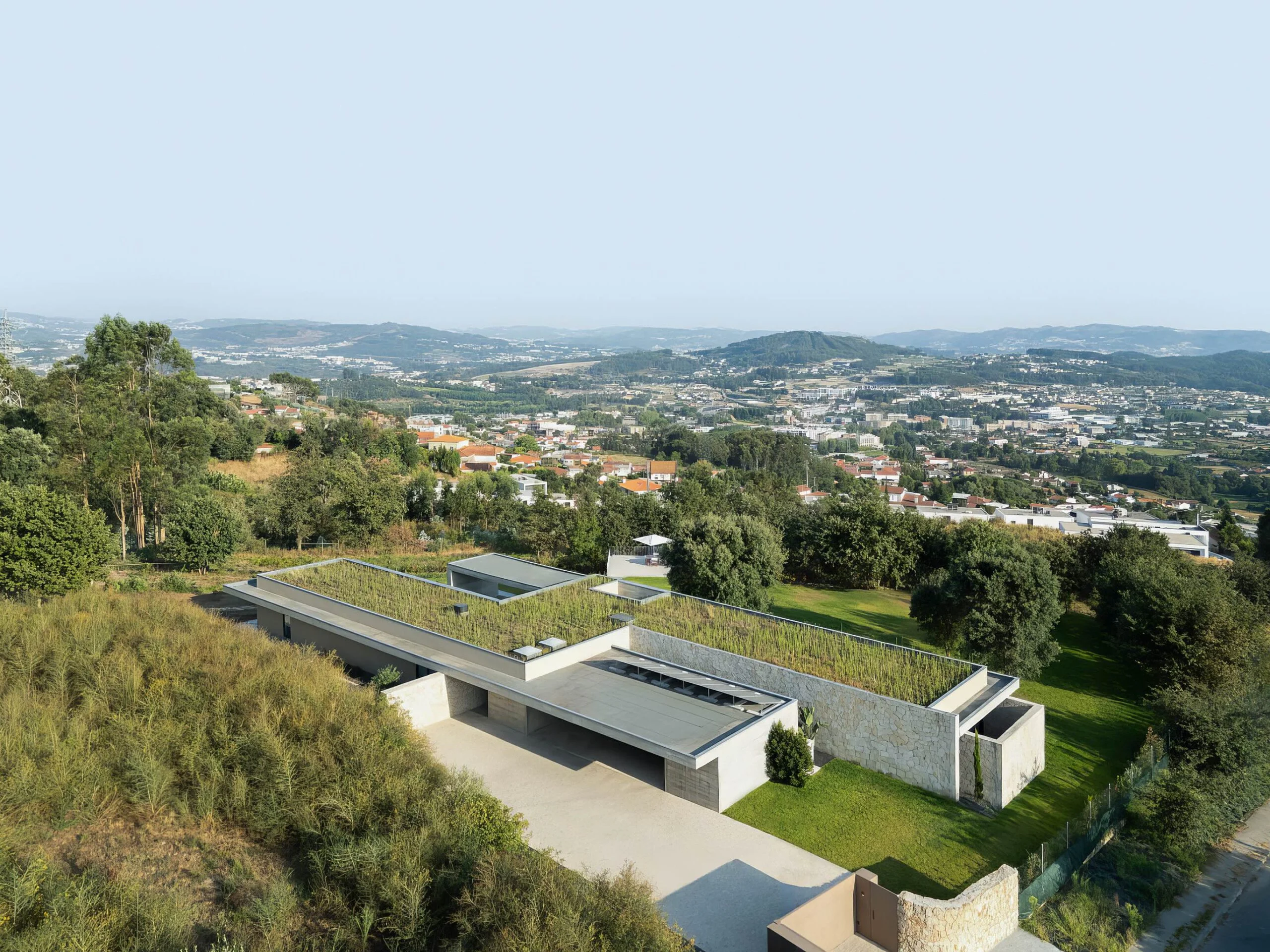 Vista aérea da moradia com cobertura ajardinada, volumes em betão aparente e alvenaria de granito, integrada na paisagem — Quinta da Casa (Habitação QC)